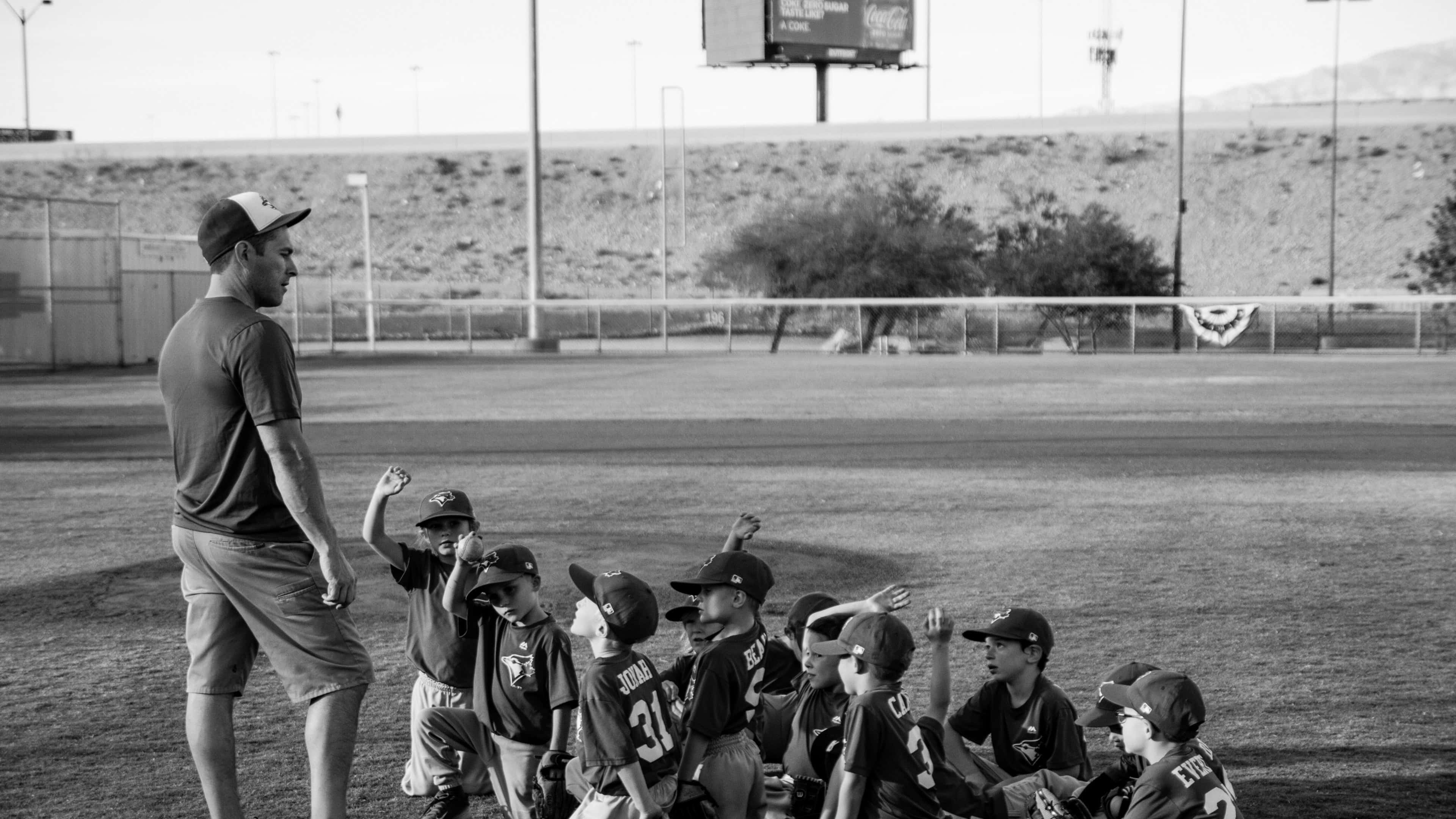 Little League Baseball 桌布 壁紙 — 一位教練在陽光明媚的球場上對一群年輕棒球選手講話，背景是沙漠山丘。