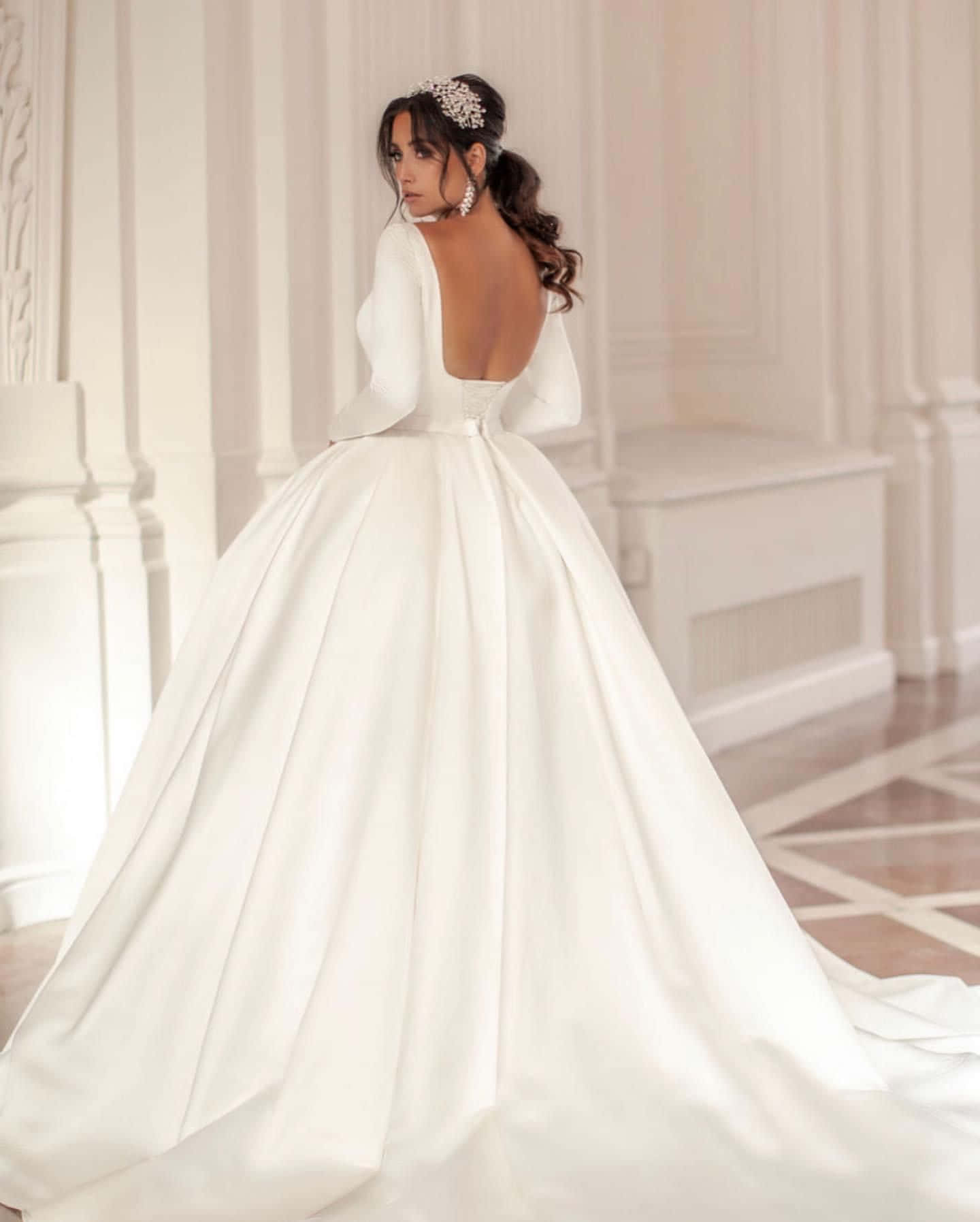 Wedding Gown 桌布 壁紙 — 一位新娘穿著寬鬆的白色緞面禮服，背部深開剪，配以珍珠飾物，站在一個優雅的白色柱廊房間中。