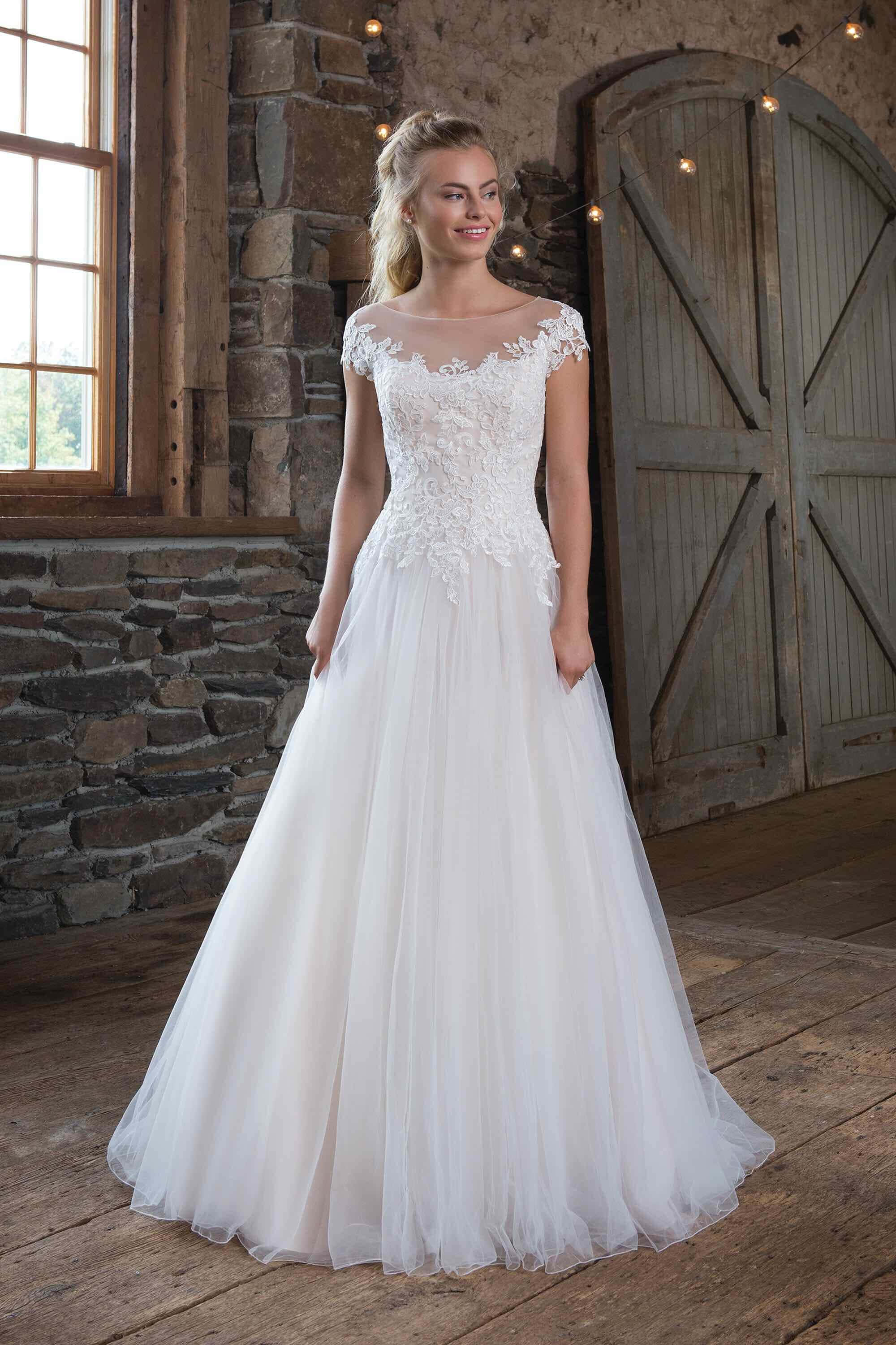 Bride's Dress 桌布 壁紙 — 一位新娘穿著白色蕾絲和薄紗禮服，在一個有石牆、木地板和大型拱門的鄉村風格房間裡微笑站立。