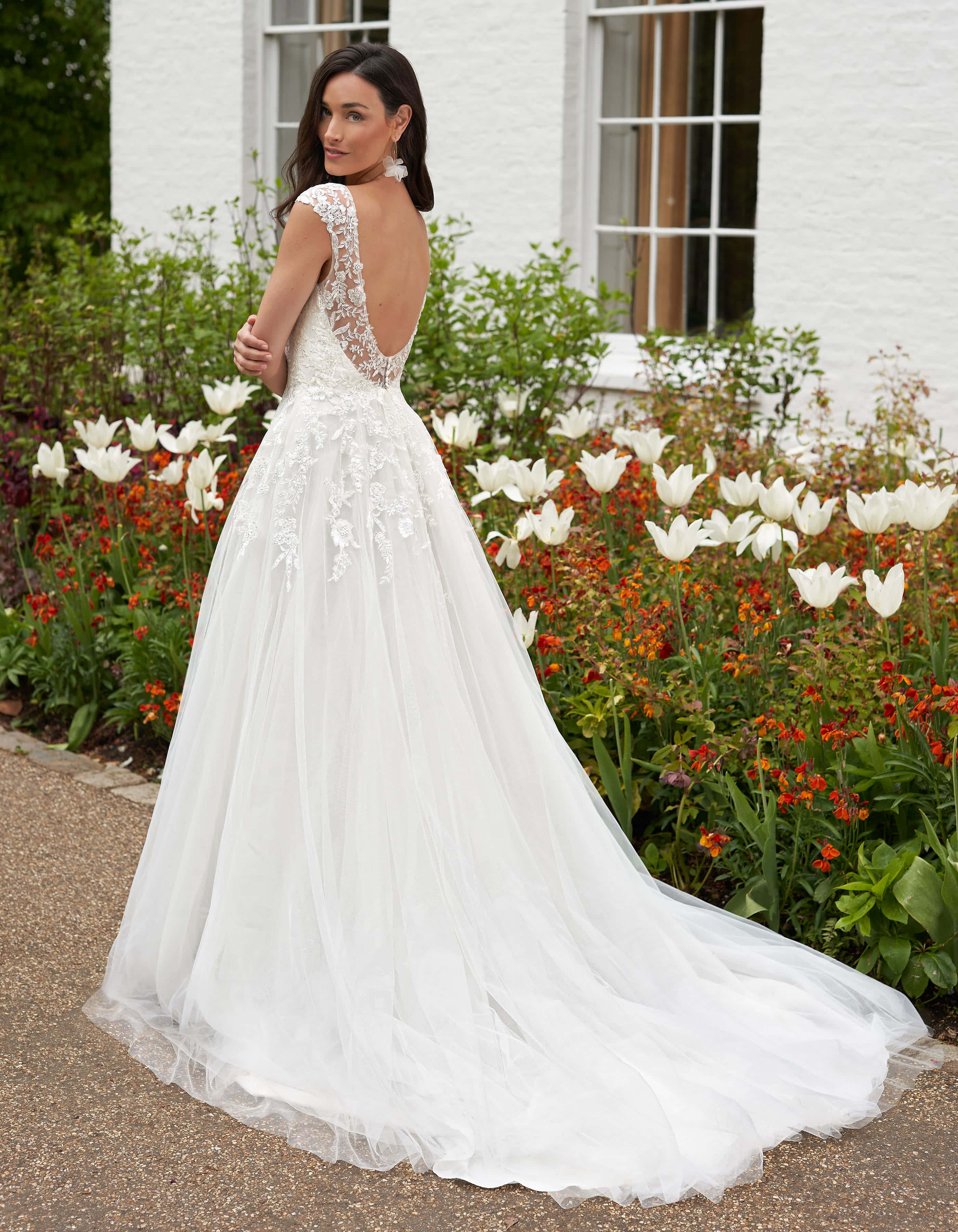 Bride's Dress 桌布 壁紙 — 一位新娘穿著白色蕾絲婚紗，露背設計，站在白色房子旁邊的白橙相間鬱金香花園中。