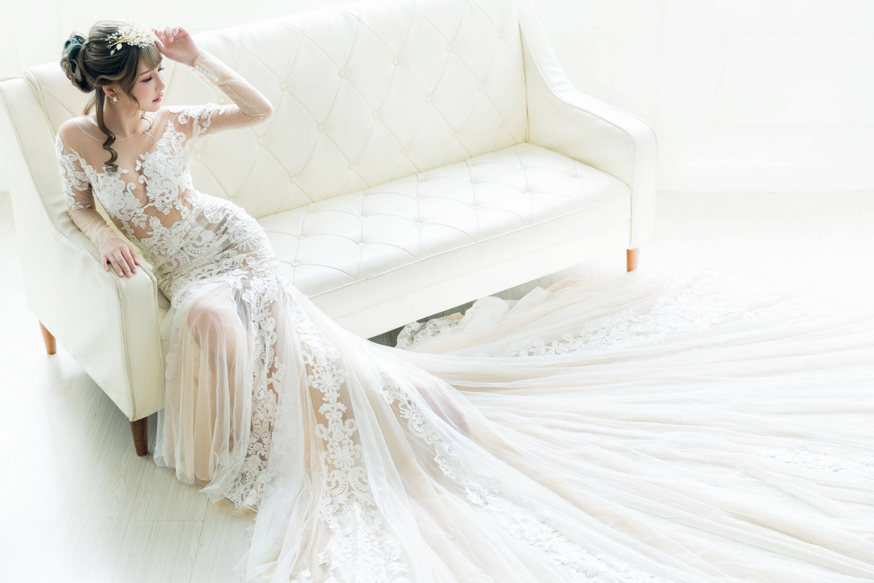 Bride 桌布 壁紙 — 一位新娘穿著精緻的白色蕾絲禮服，優雅地坐在明亮簡約房間裡的簇絨白色沙發上。