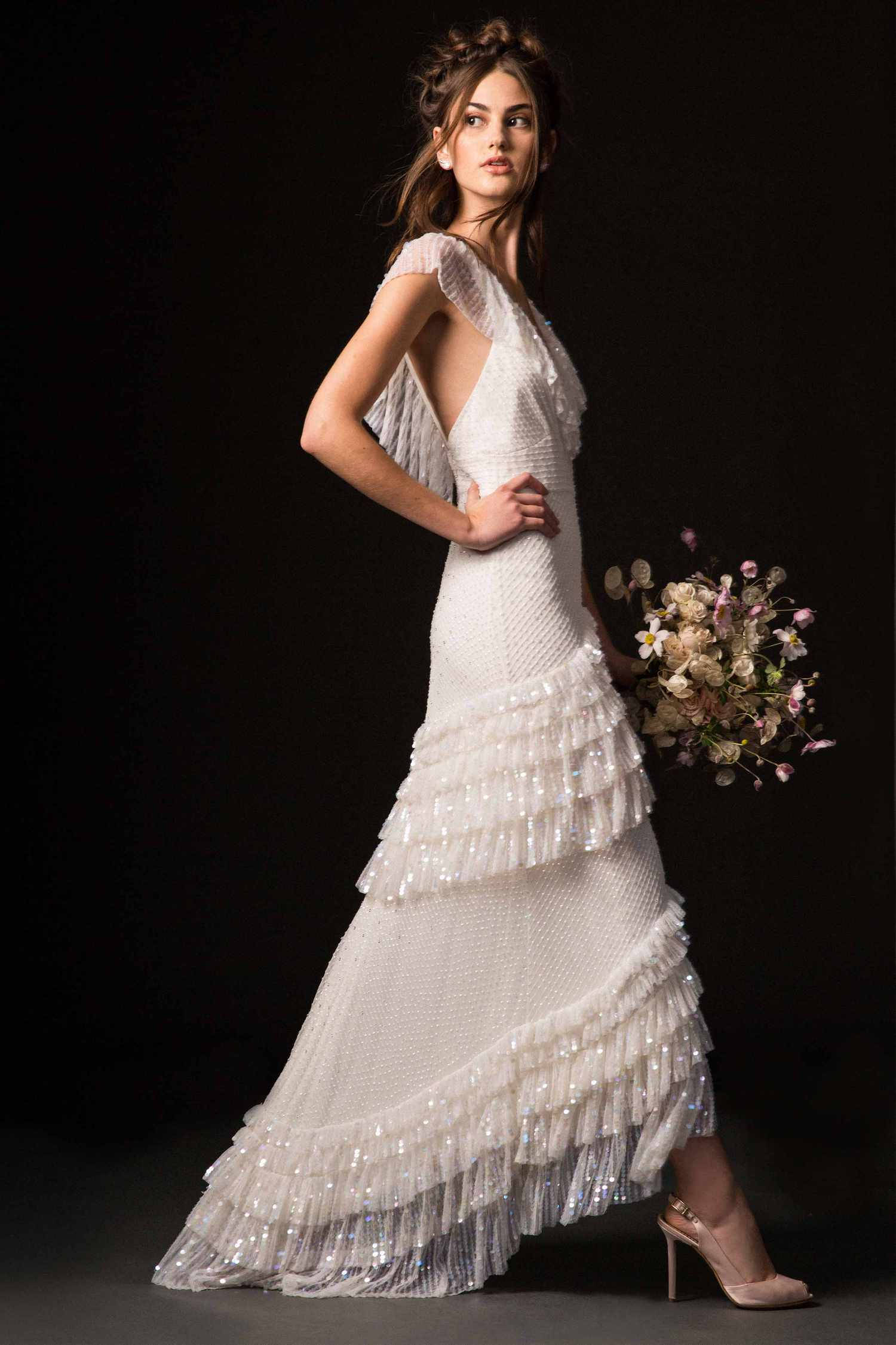 Bride 桌布 壁紙 — 一位新娘穿著質感豐富的白色禮服，帶有層疊荷葉邊和亮片，在黑色背景前自信地擺姿勢，手持一束柔和粉彩花束。