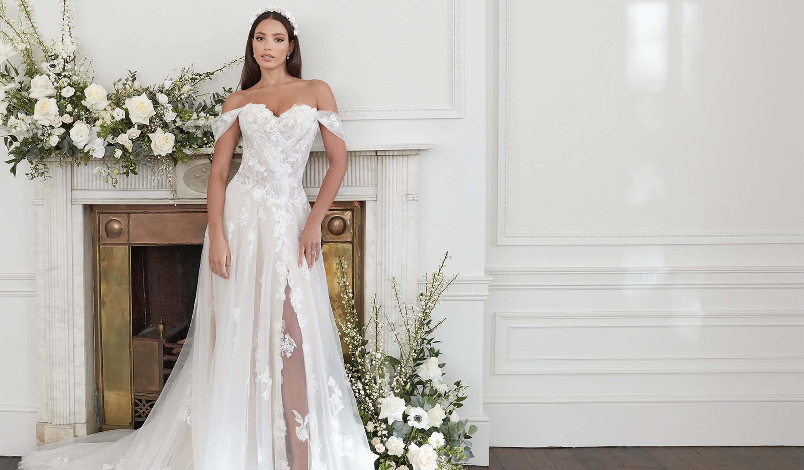 Bride's Dress 桌布 壁紙 — 一位新娘穿著露肩白色蕾絲婚紗，優雅地站在一個裝飾著白色花卉佈置的大理石壁爐旁。