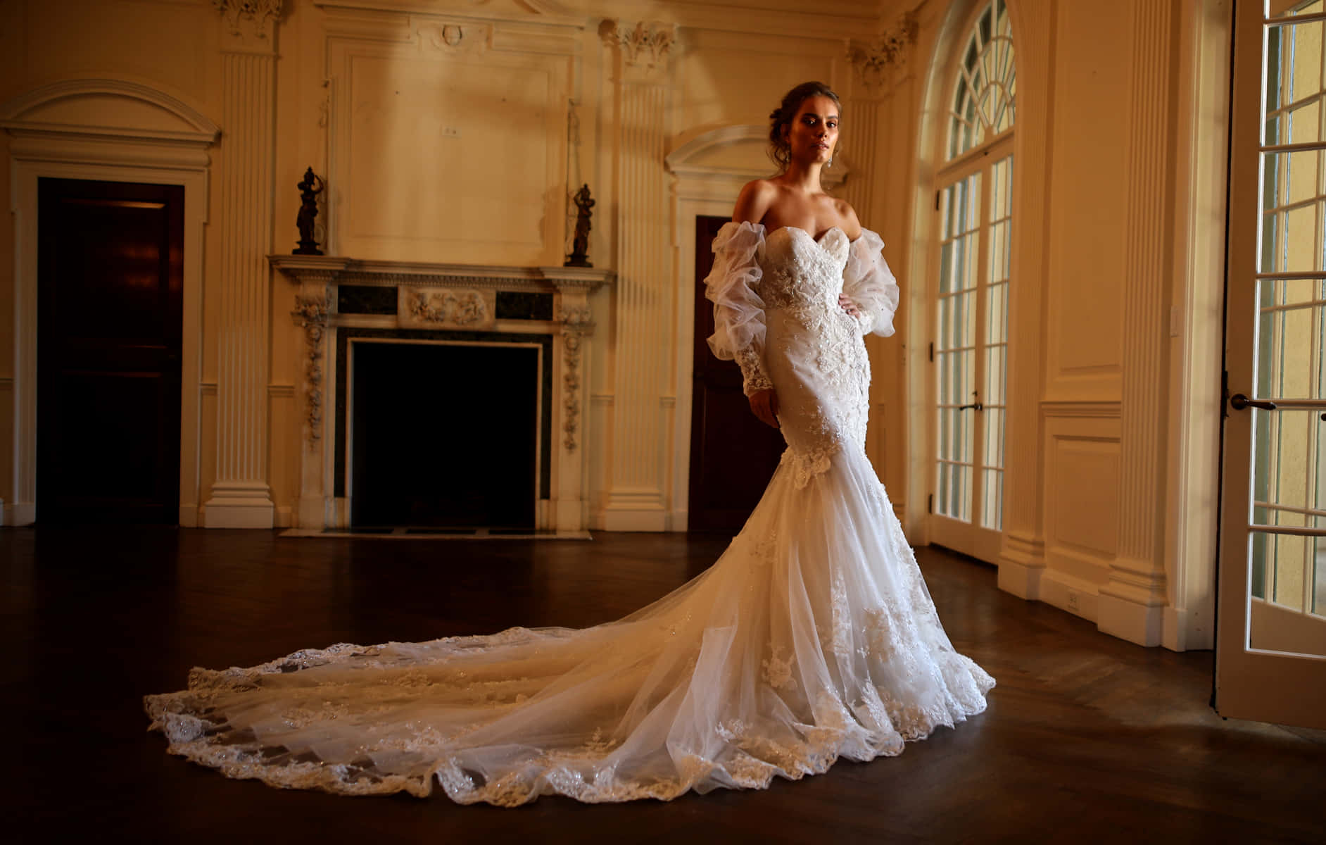 Bride's Dress 桌布 壁紙 — 一位新娘穿著露肩蕾絲美人魚婚紗，優雅地站在一個宏偉、陽光充足的維多利亞式大廳中，大廳有華麗的壁爐和高大的窗戶。