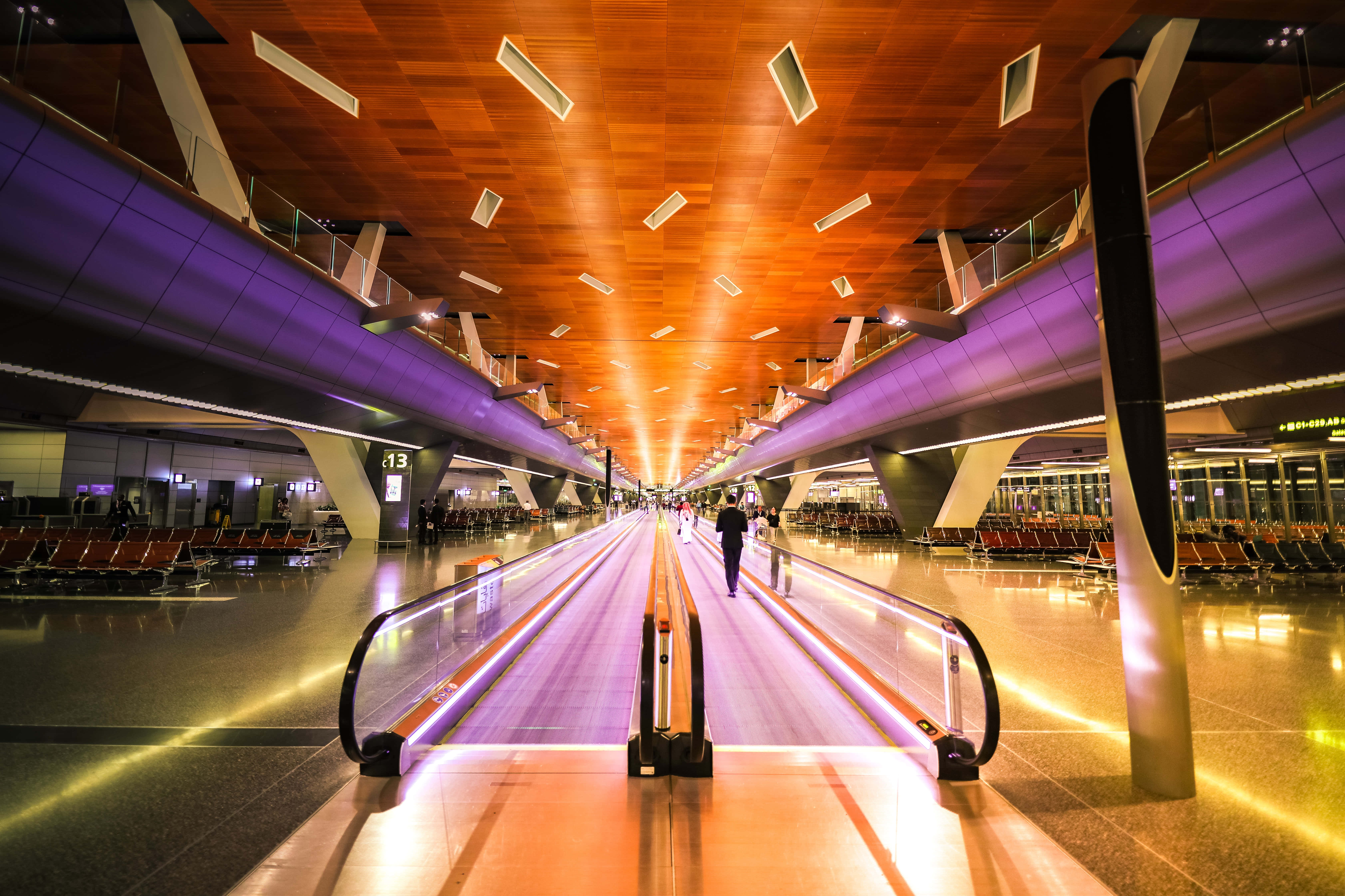 Airport 桌布 壁紙 — 一位旅客在現代機場航廈內，沿著發光的紫色自動人行道行走，天花板採用溫暖的木質設計，並配以環境照明。