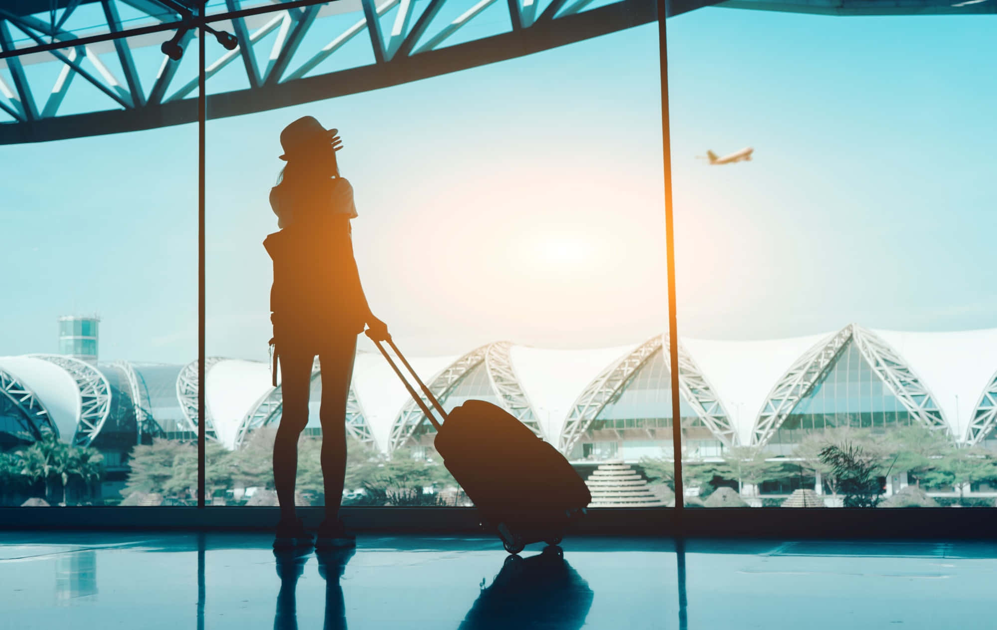 Traveling 桌布 壁紙 — 一位旅客的剪影，帶著行李箱站在玻璃牆旁，看著飛機在日落時分飛過現代化的機場航廈。