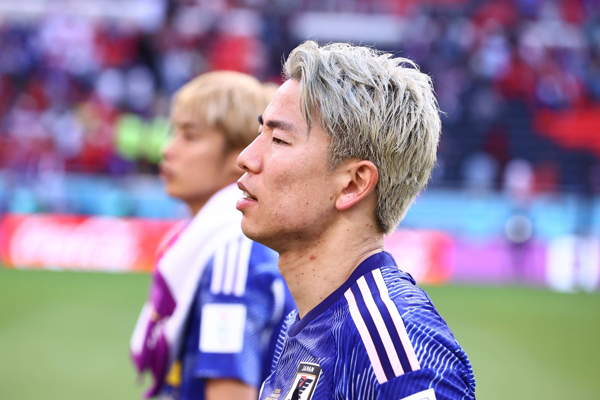 Football Players 桌布 壁紙 — 一位日本足球員的近距離特寫，銀色頭髮，身穿藍色球衣，站在綠色球場上，背景是模糊的體育場人群。