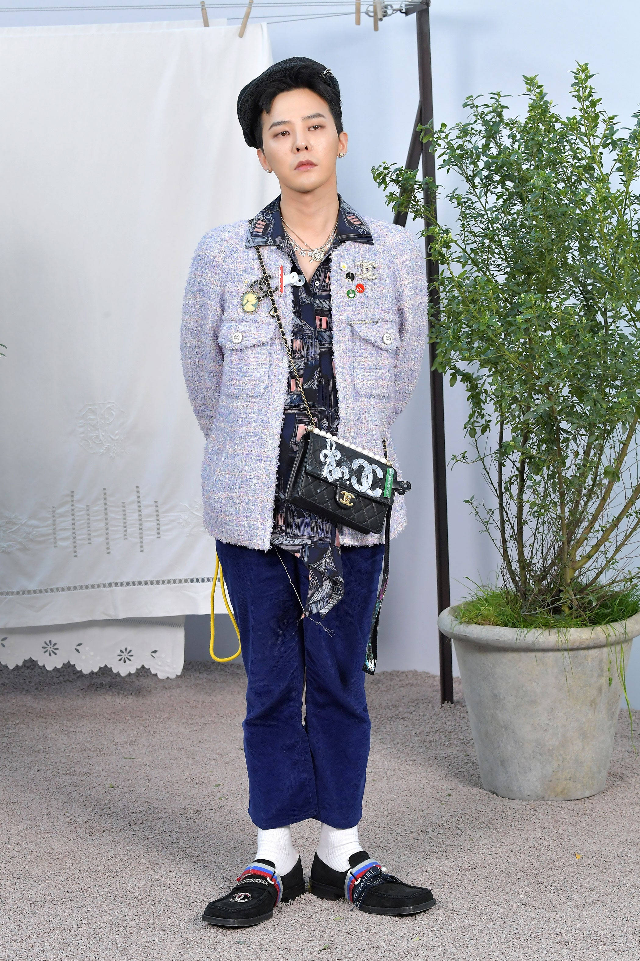 G-dragon 桌布 壁紙 — 一位時尚人士穿著質感西裝外套和藍色褲子，自信地站在盆栽旁邊，柔和日光下。