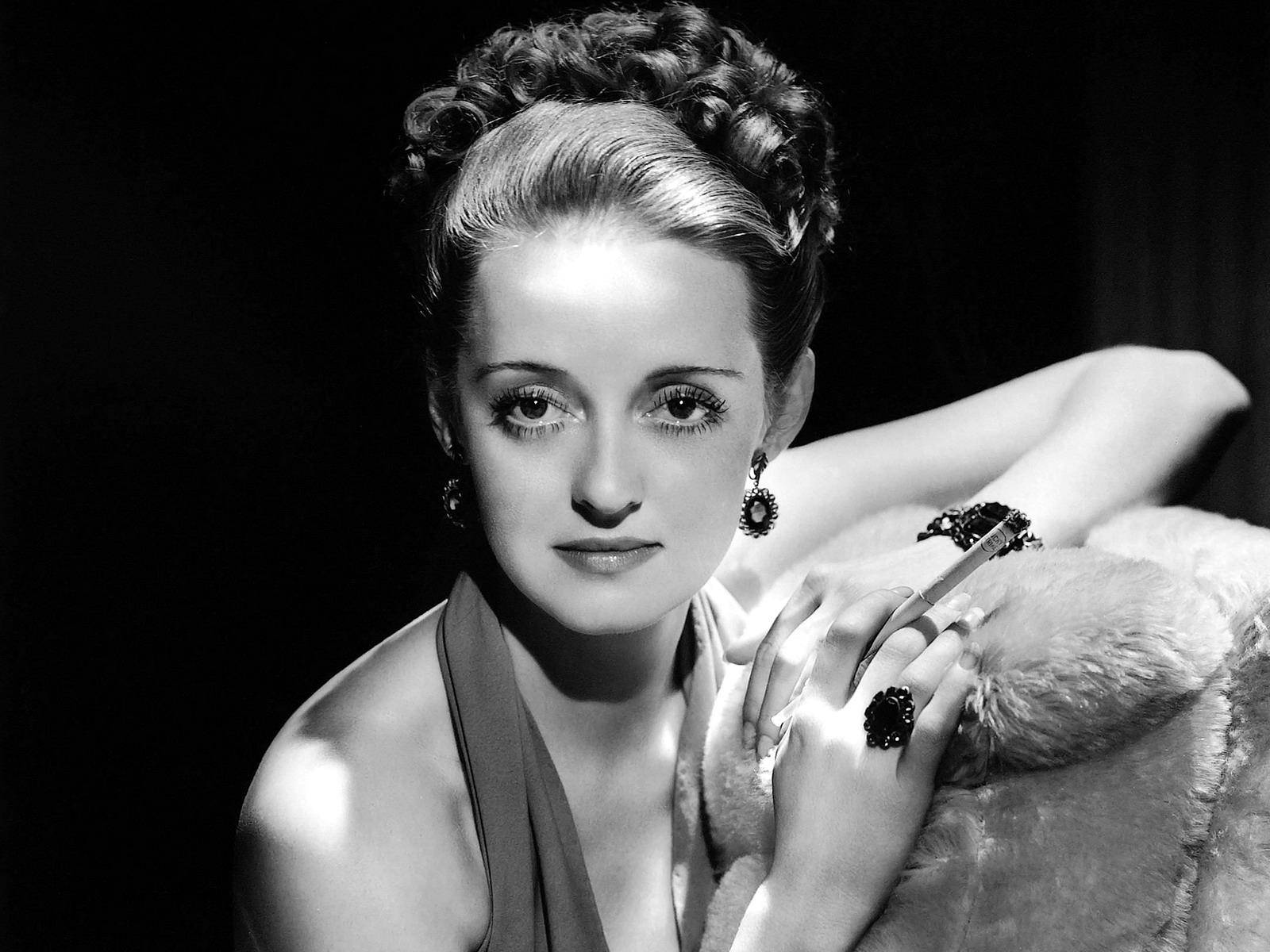 Bette Davis 桌布 壁紙 — 一位時尚女性，捲髮配戴大膽耳環，披著皮草披肩，在深色背景下優雅擺姿勢。