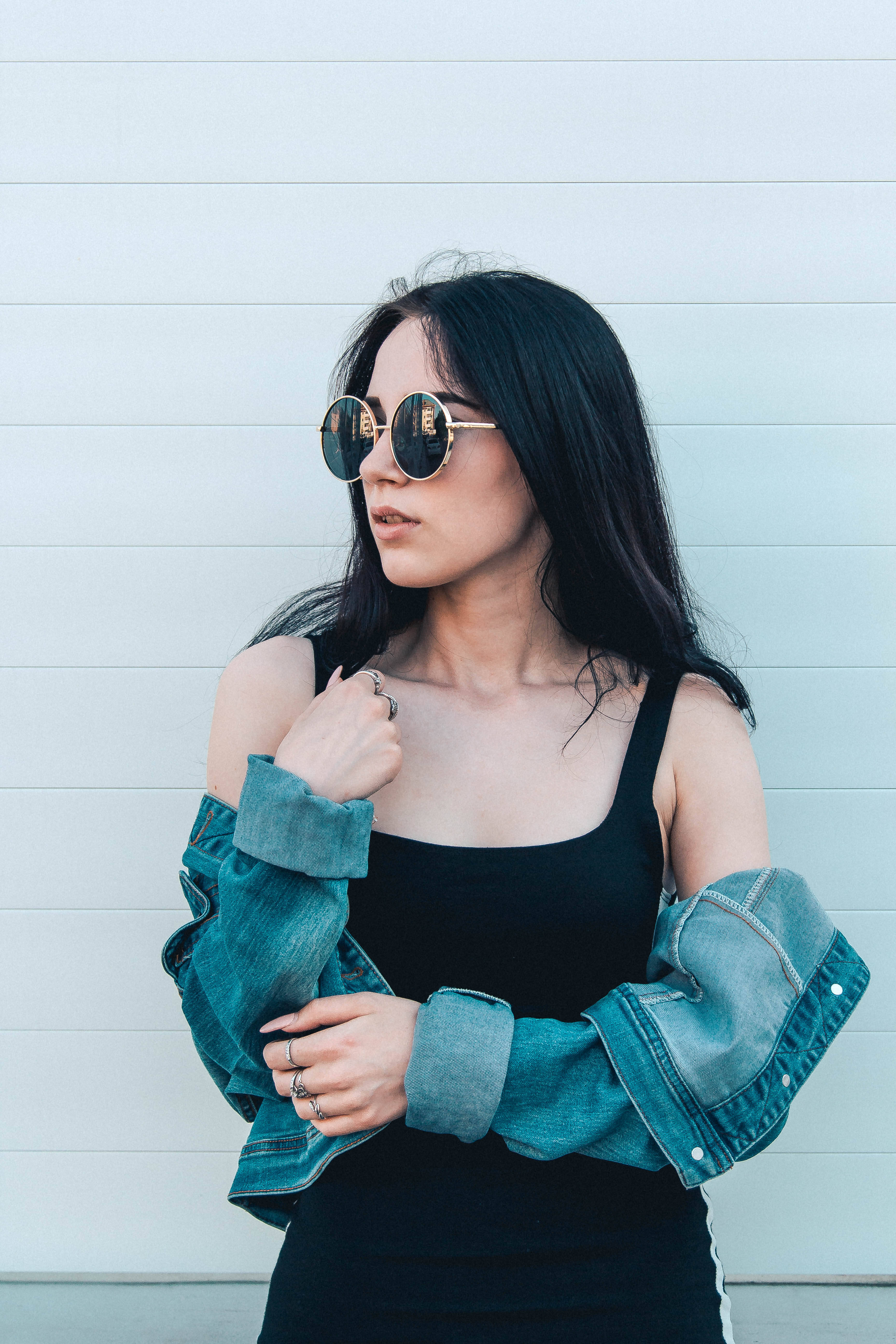 Trendy 桌布 壁紙 — 一位時尚女性，深色頭髮，戴著圓形太陽眼鏡，穿著黑色上衣和牛仔外套，自信地靠在淺灰色鑲板牆上擺姿勢。