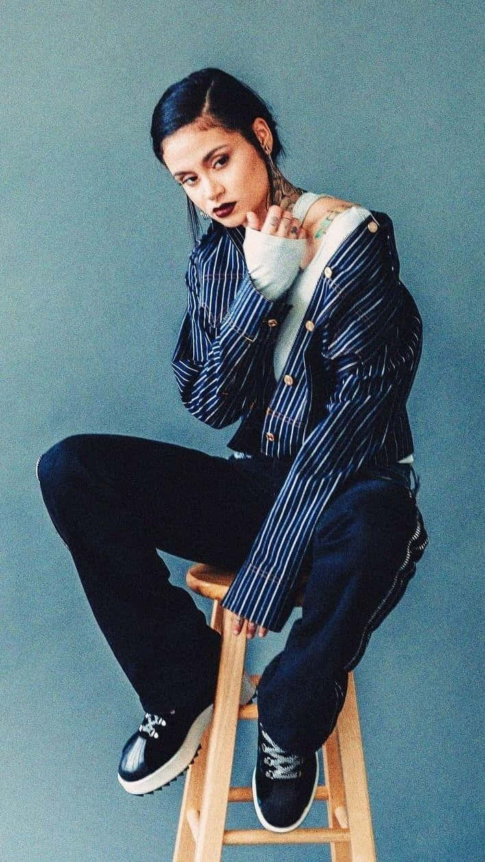 kehlani 桌布 壁紙 — 一位時尚女性，深色頭髮和刺青，自信地坐在木凳上，背景是柔和的藍色牆壁，穿著細條紋外套和黑色褲子。