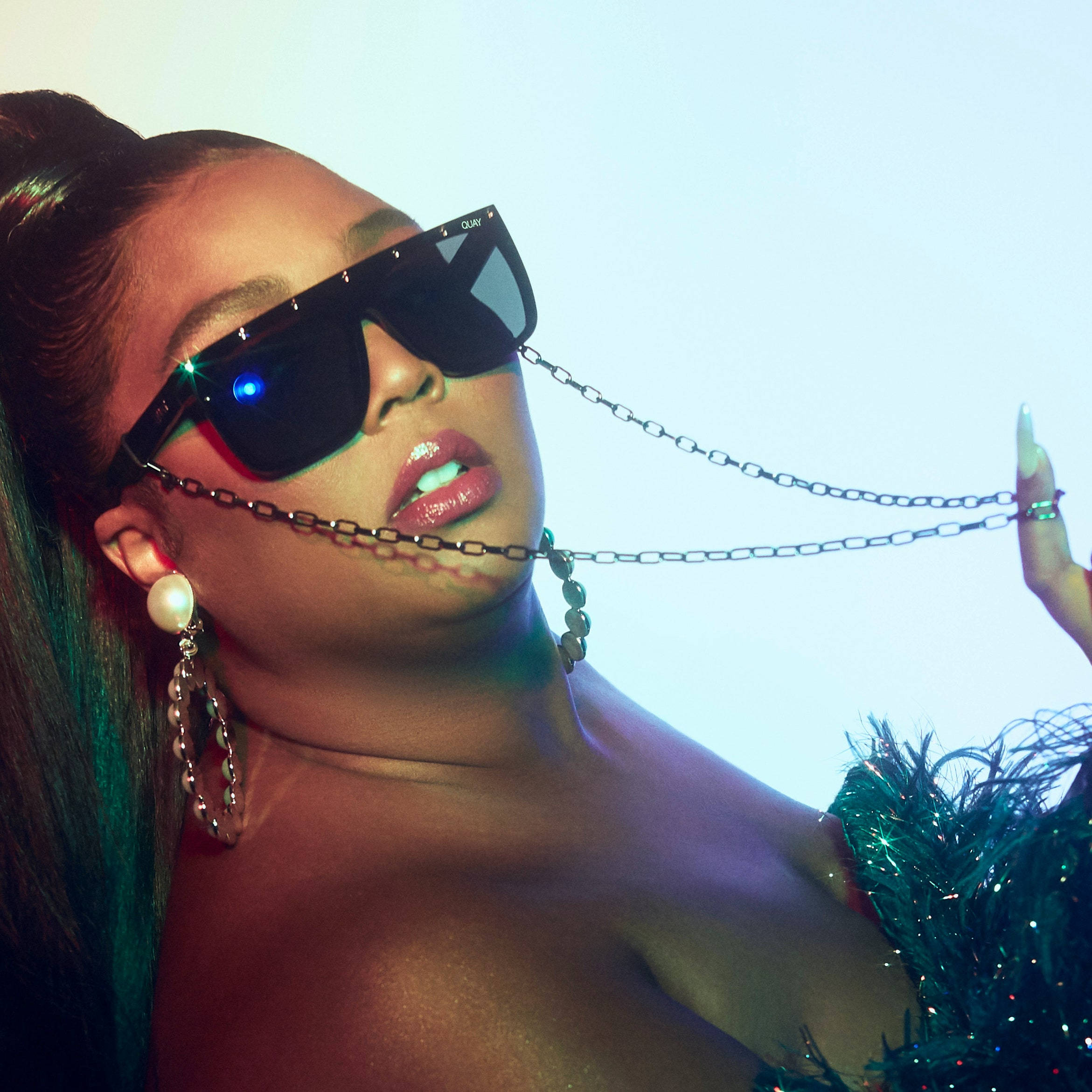 Lizzo 桌布 壁紙 — 一位時尚女性戴著大膽的太陽眼鏡和鏈條飾品，在發光的藍色背景前擺姿勢。
