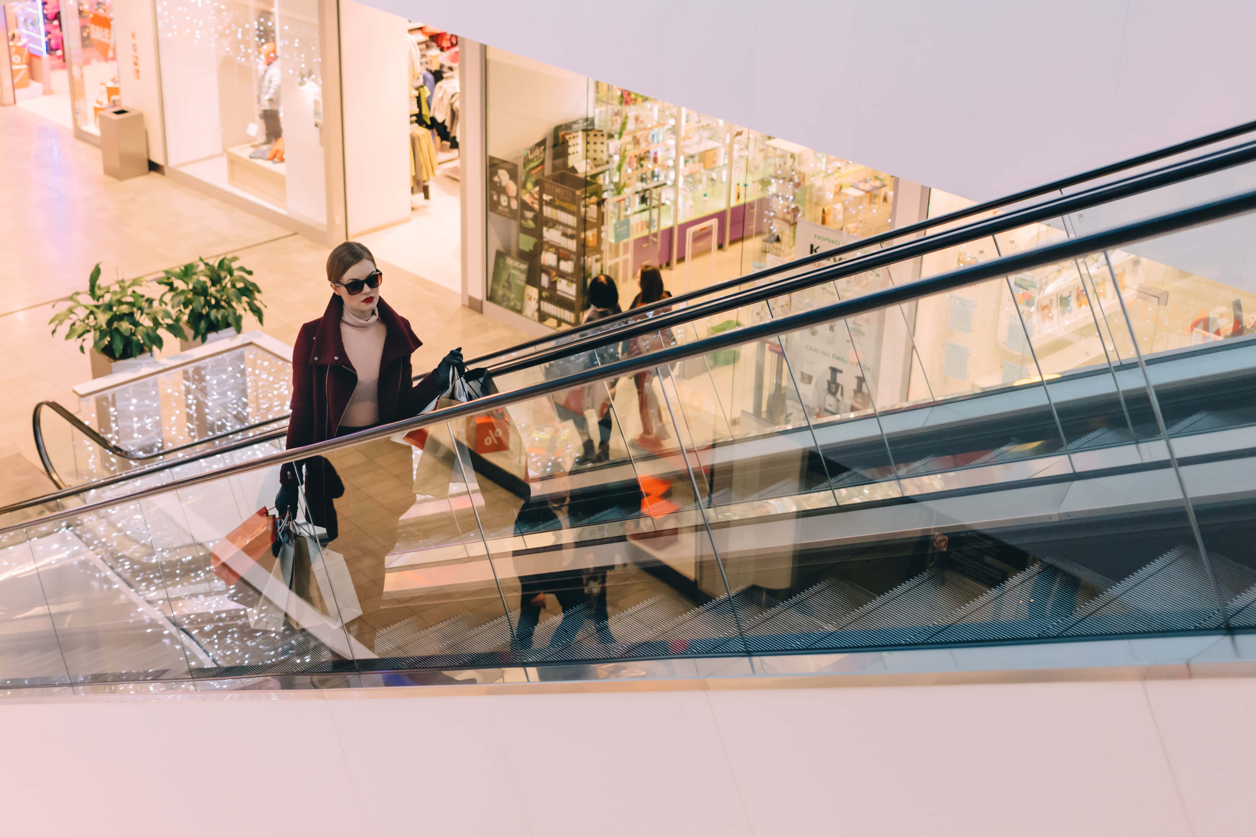 Sunglasses 桌布 壁紙 — 一位時尚女性戴著太陽眼鏡，在光線明亮的購物中心內乘坐玻璃圍封的扶手電梯，手持購物袋。