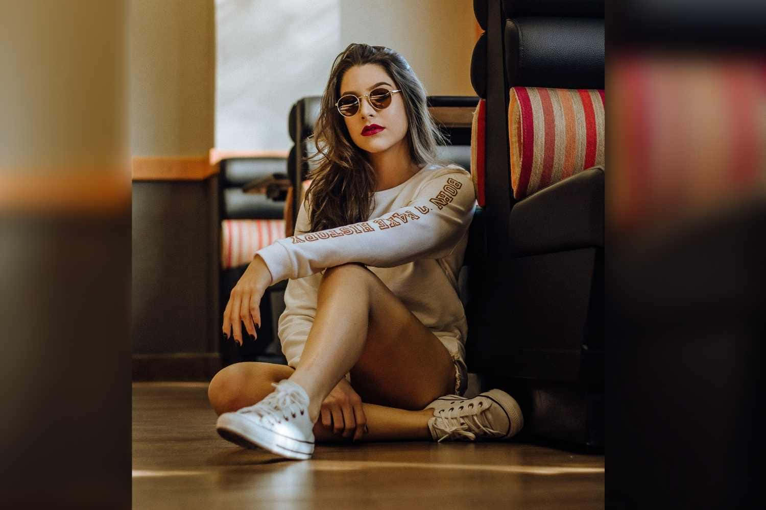 美女 桌布 壁紙 — 一位時尚女性戴著太陽眼鏡，在溫暖照明的室內空間中，盤腿坐在木地板上，旁邊是一張條紋扶手椅。