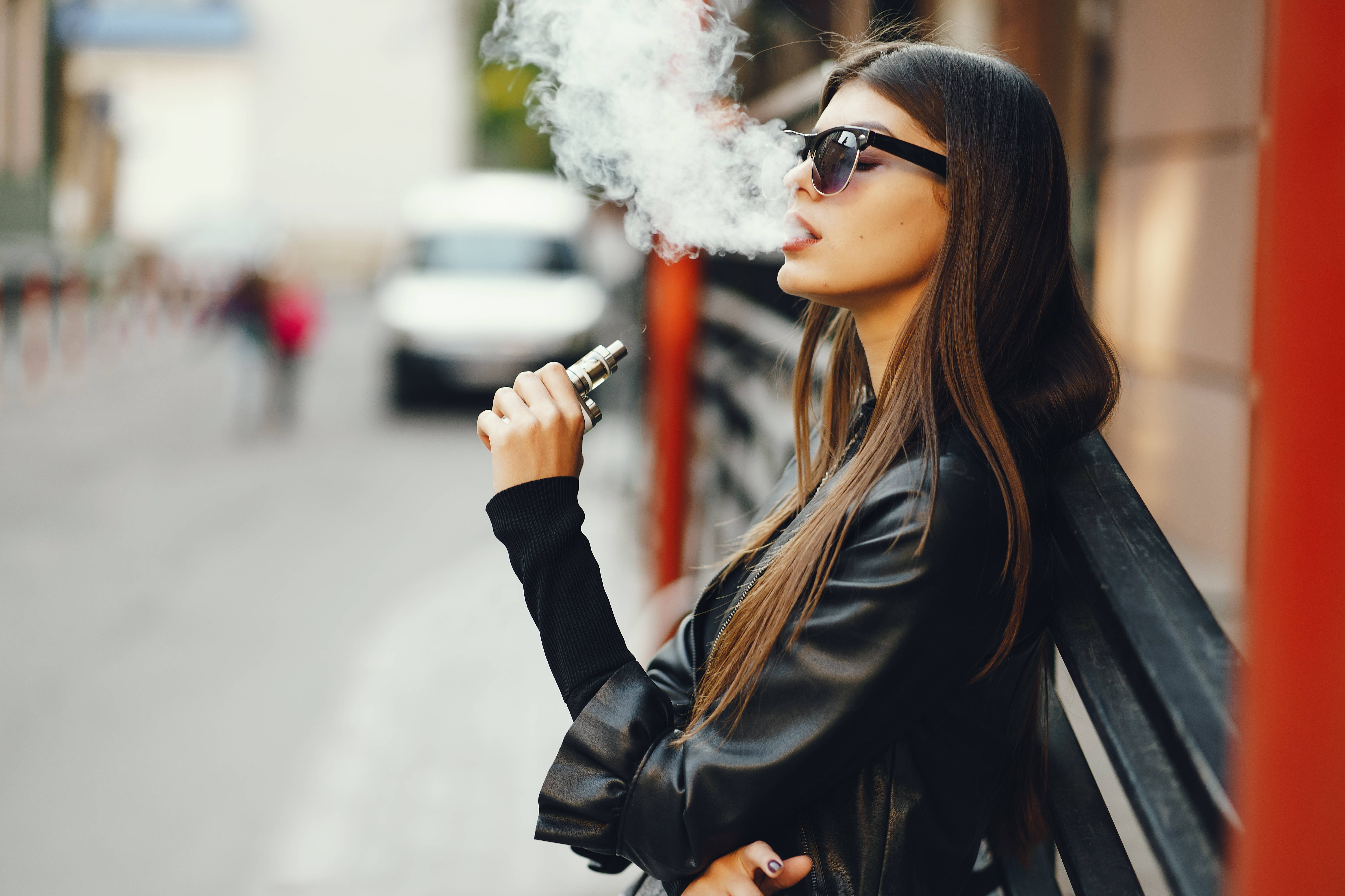 抽菸 桌布 壁紙 — 一位時尚女性戴著太陽眼鏡和黑色皮外套，在陽光明媚的城市街道上靠著長椅呼出蒸氣。