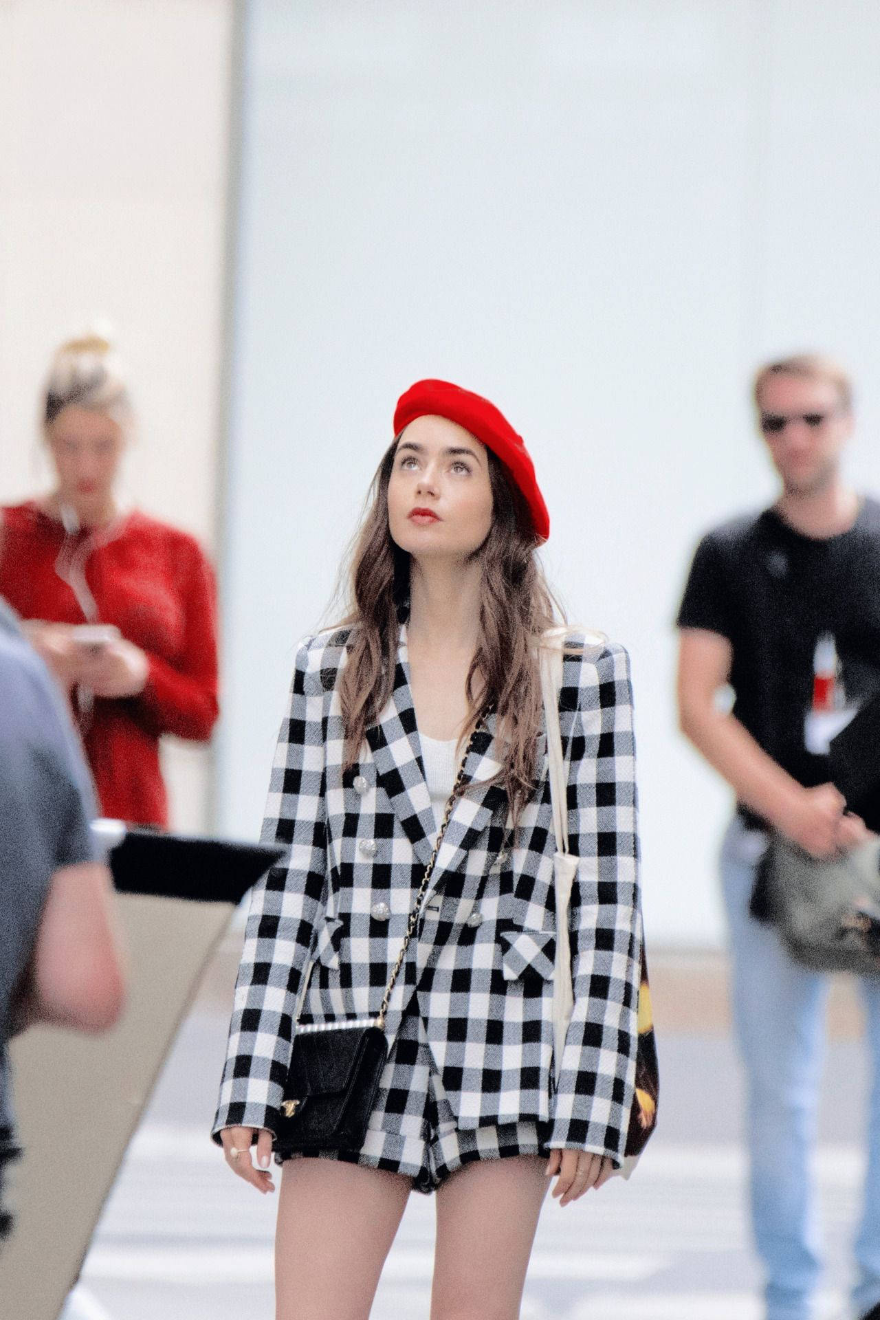 Emily In Paris 桌布 壁紙 — 一位時尚女性戴著紅色貝雷帽，身穿黑白格紋西裝外套，自信地走過明亮的城市環境。