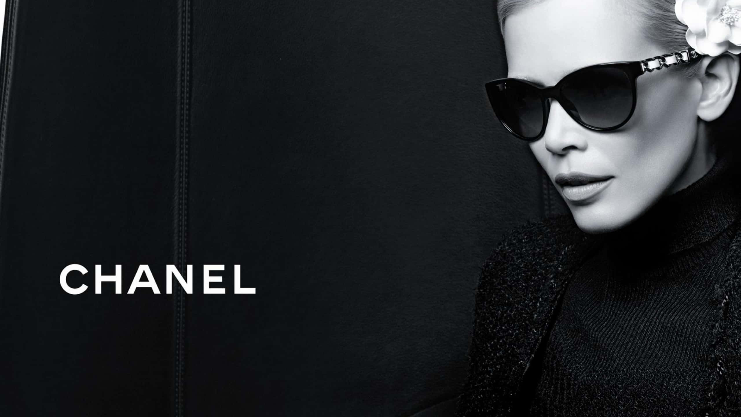 Chanel 桌布 壁紙 — 一位時尚女性戴著黑色太陽眼鏡和質感外套，在帶有 Chanel 標誌的深色背景前擺姿勢。