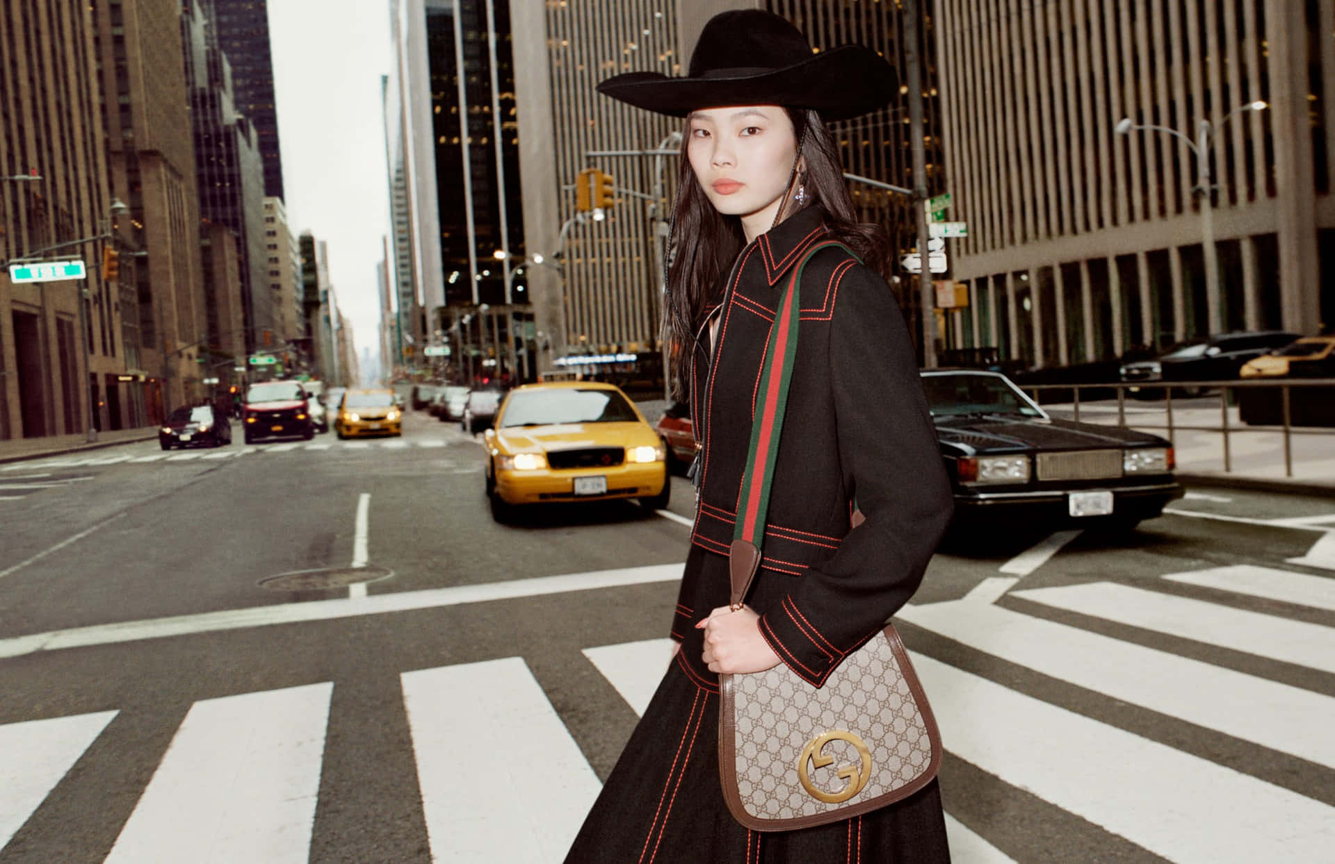 Gucci 桌布 壁紙 — 一位時尚女性戴著黑色寬邊帽和外套，穿過繁忙的城市街道，背景有黃色計程車和高樓大廈。