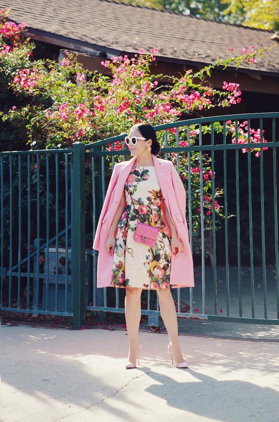 Pink Coat 桌布 壁紙 — 一位時尚女性穿著碎花裙和粉紅色外套，自信地站在一個裝飾著盛開粉紅色花朵的綠色籬笆前。