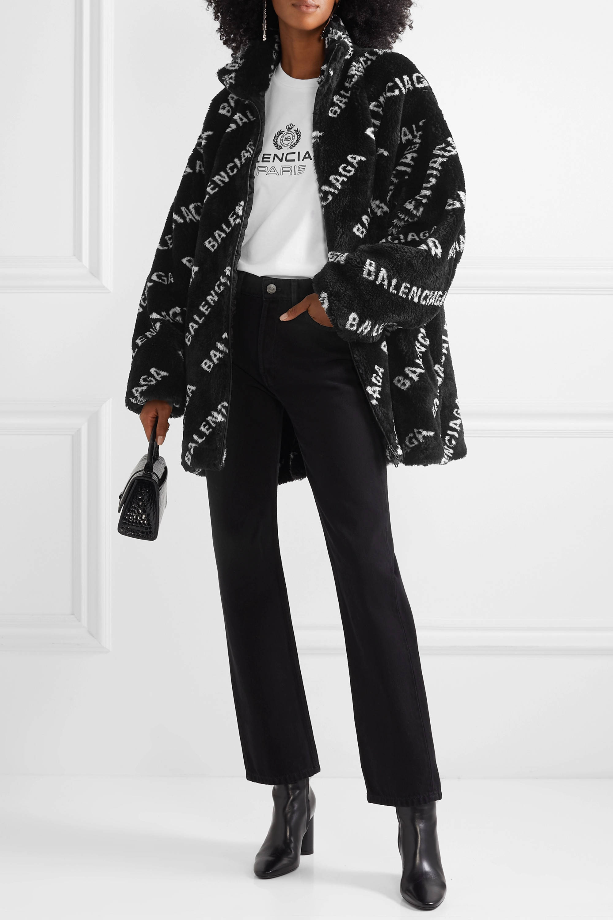 Balenciaga 桌布 壁紙 — 一位時尚女性穿著黑色Balenciaga標誌印花泰迪外套，內搭白色T恤和黑色褲子，背景是簡約的白色牆壁。
