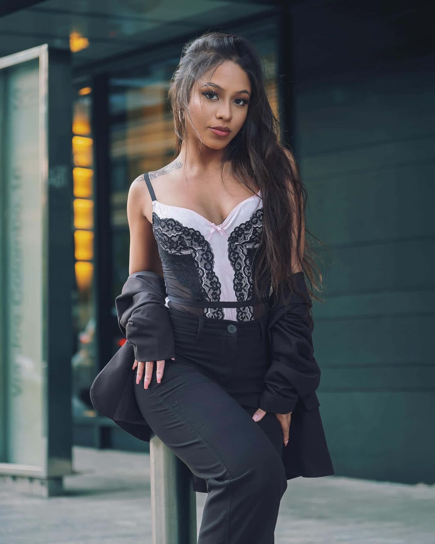 Single Latino Woman 壁紙 — 一位時尚女性穿著黑色蕾絲上衣和西裝外套，在黃昏時自信地靠在一棟現代玻璃建築上。