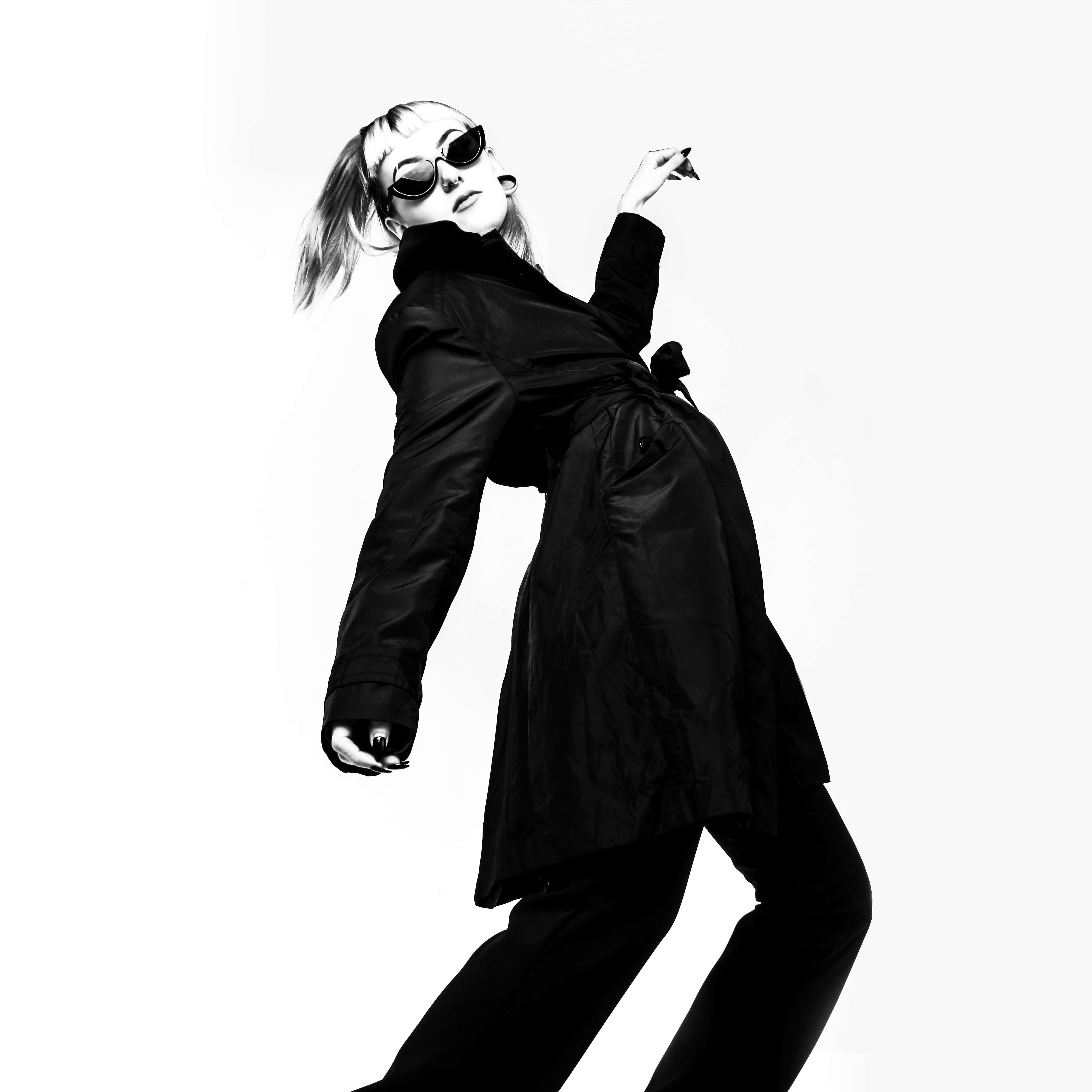 Black And White Fashion 桌布 壁紙 — 一位時尚女性穿著黑色風衣和太陽眼鏡，在純白背景下擺出動感姿勢。