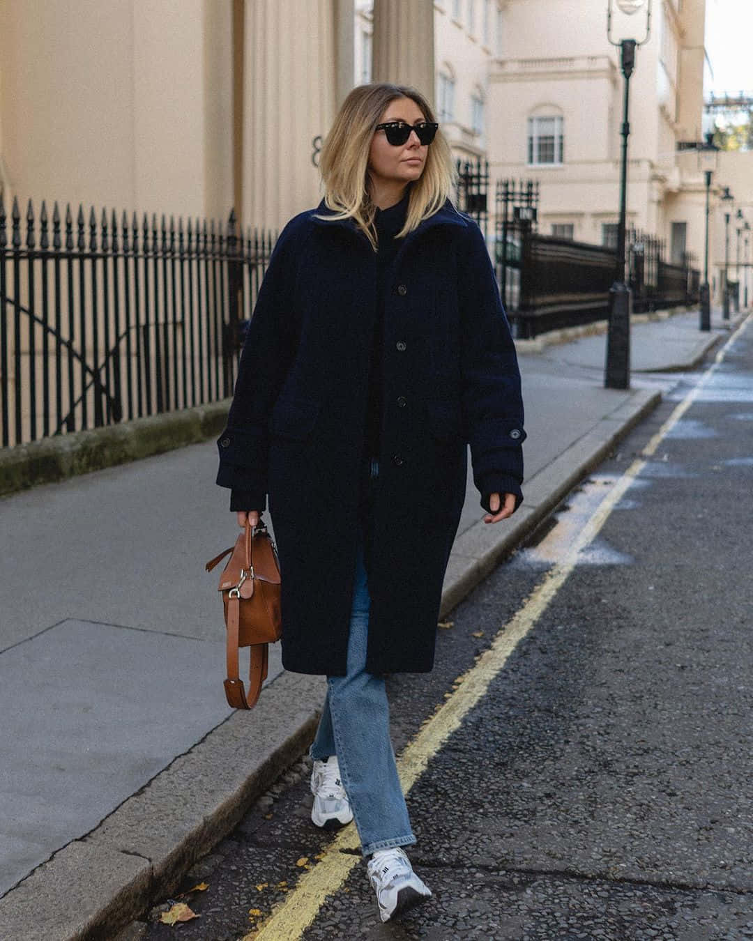 Navy Blue Coat 桌布 壁紙 — 一位時尚女性身穿海軍藍外套和太陽眼鏡，自信地走在濕漉漉的城市街道上，兩旁是米色建築和黑色圍欄。
