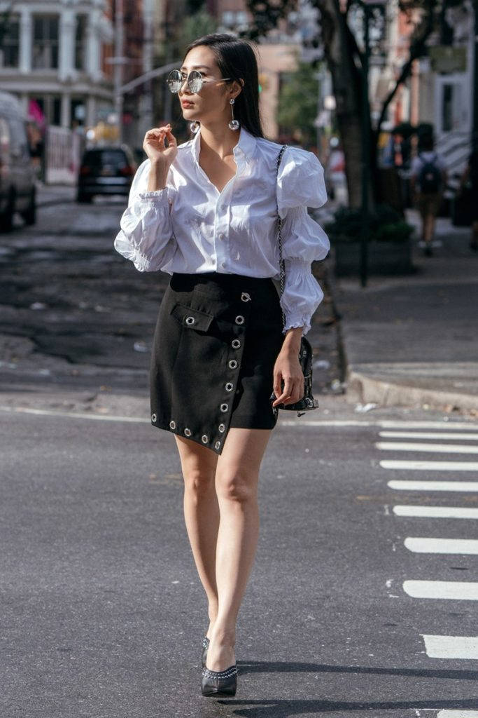 Trendy Aesthetic 桌布 壁紙 — 一位時尚女性身穿白色襯衫和黑色裙子，戴著太陽眼鏡，手提一個小包，自信地走過陽光普照的城市街道。