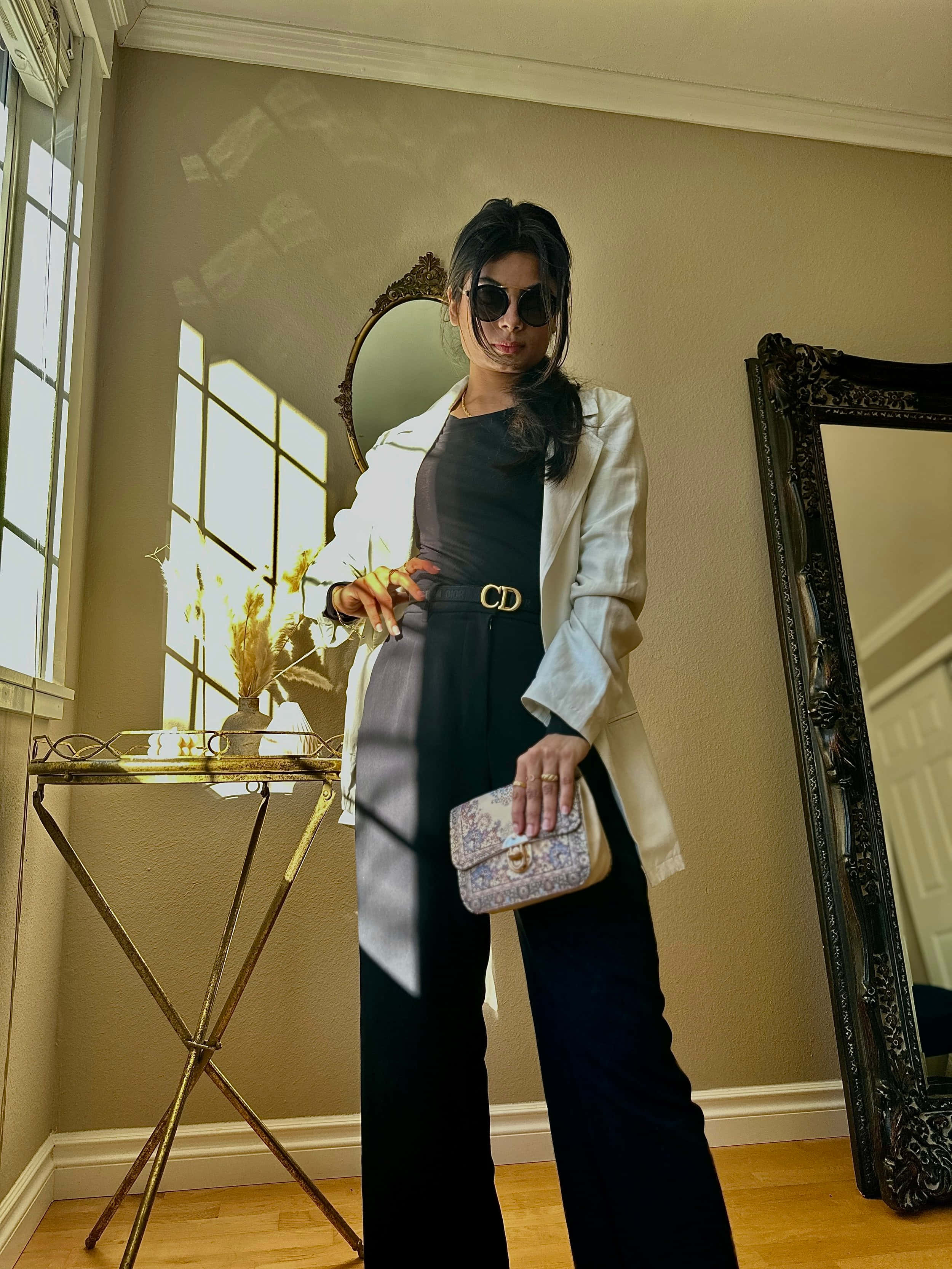 Minimalist Fashion 桌布 壁紙 — 一位時尚女性身穿白色西裝外套和黑色長褲，在陽光充足的房間裡自信地擺姿勢，房間內有華麗的鏡子和一張復古邊桌。