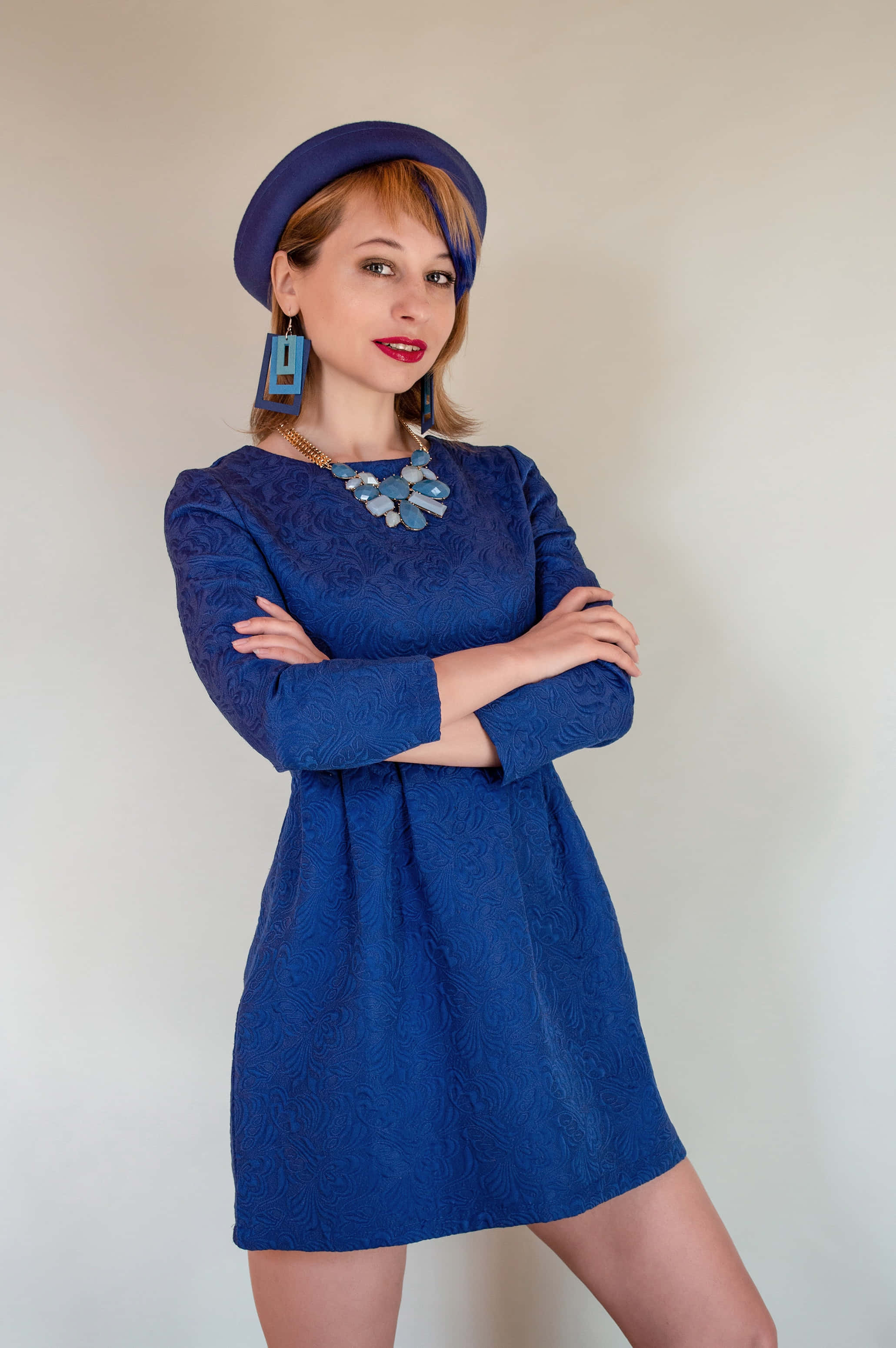 Navy Blue Dress 桌布 壁紙 — 一位時尚女性身穿皇家藍色質感連衣裙和配套帽子，在純色背景前自信地交叉雙臂擺姿勢。
