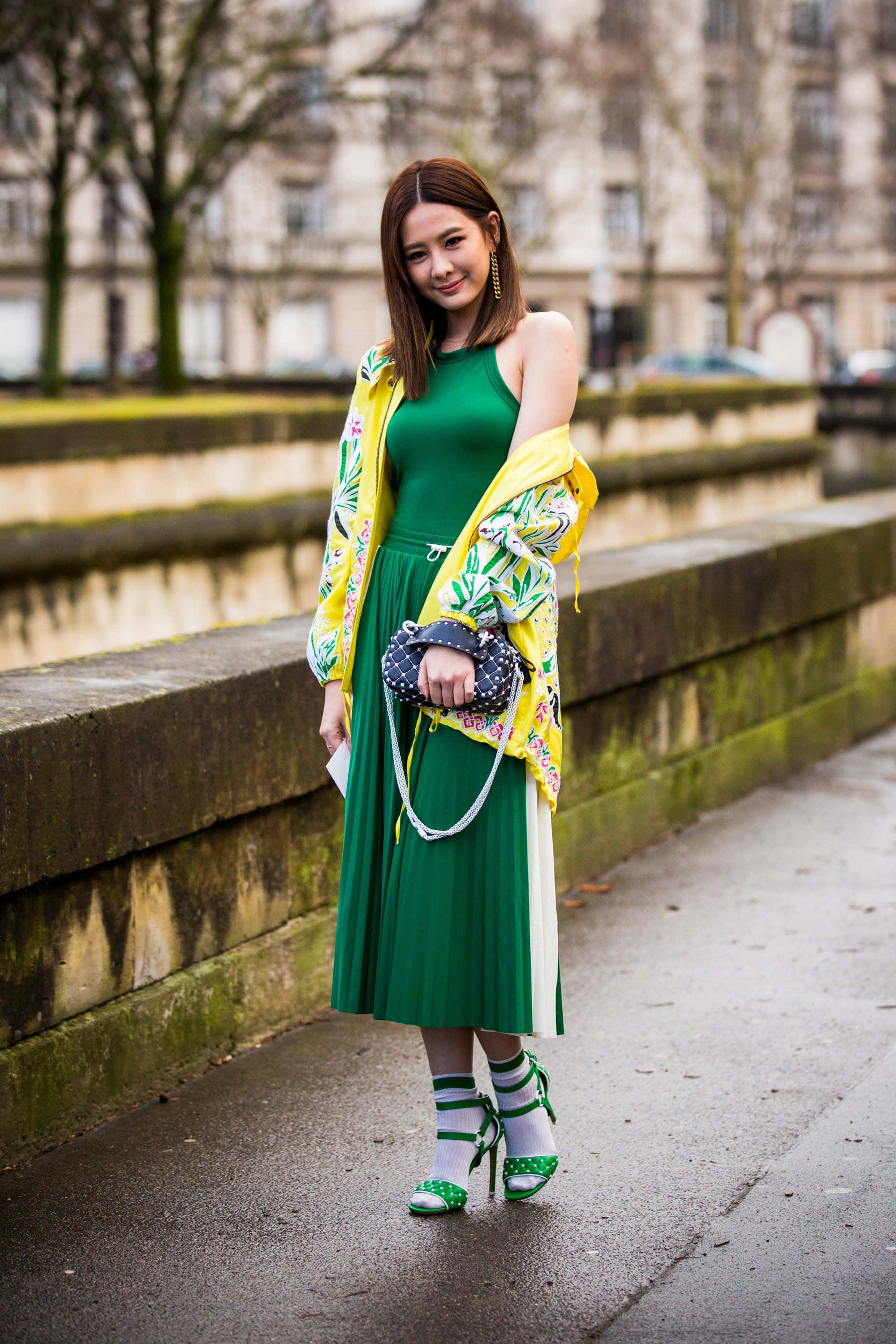 Green Fashion 桌布 壁紙 — 一位時尚女性身穿鮮綠色連衣裙和花卉黃色外套，在運河附近潮濕的城市人行道上自信地擺姿勢。