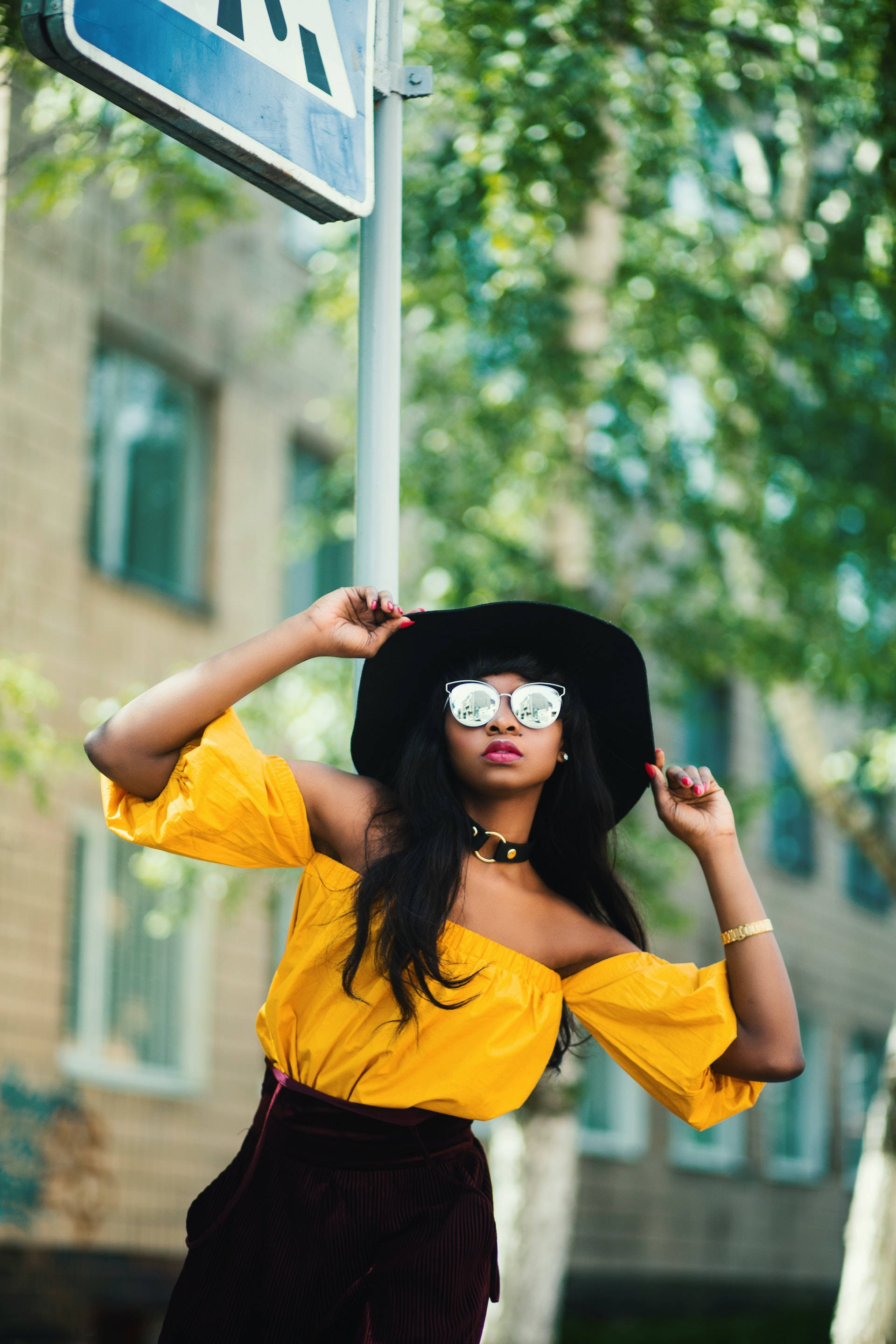 Black Girl 桌布 壁紙 — 一位時尚女性身穿黃色上衣和黑色帽子，在路牌下自信地擺姿勢，身後有茂密的綠樹。