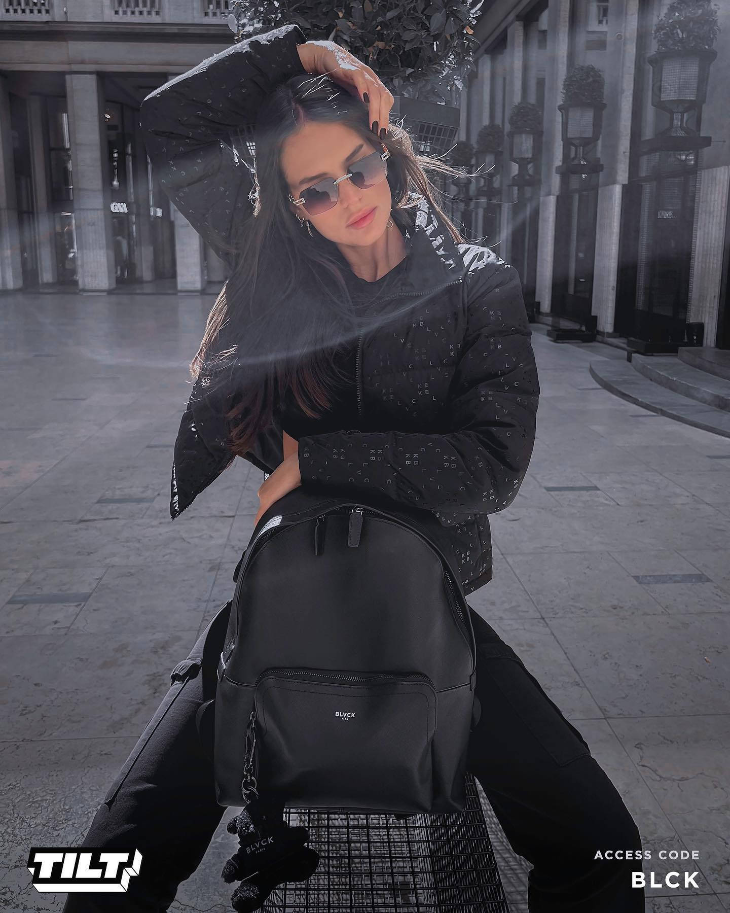 Blvck Paris 桌布 壁紙 — 一位時尚女性身穿黑色圖案外套和太陽眼鏡，自信地在城市廣場上擺姿勢，背著黑色背包。
