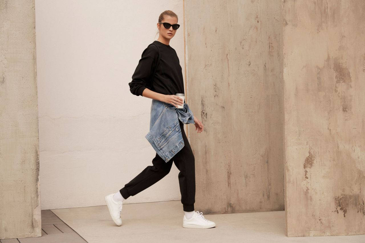 Sunglasses 桌布 壁紙 — 一位時尚女性身穿黑色服裝和白色運動鞋，自信地走在紋理混凝土牆之間，手持咖啡杯。