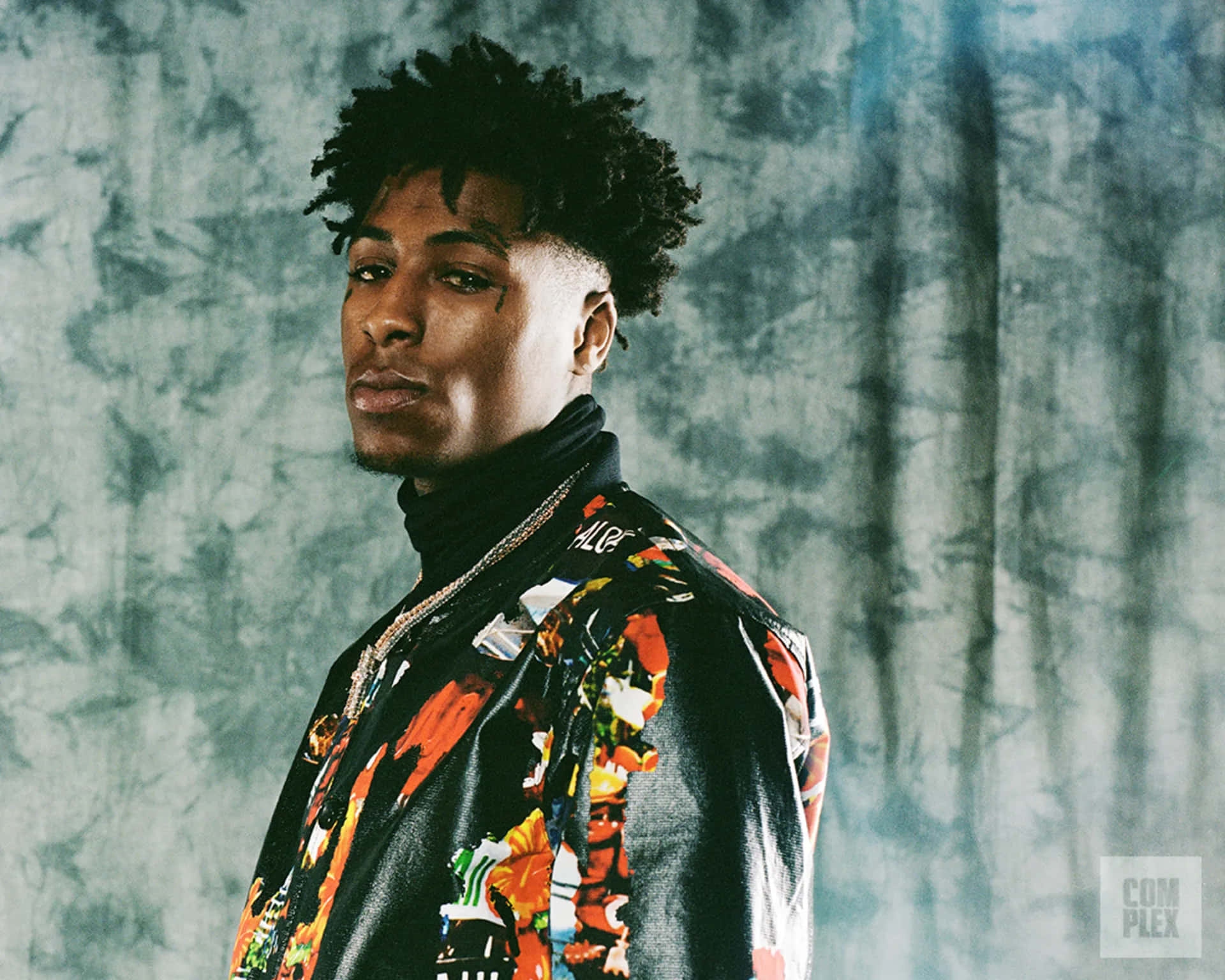 Youngboy 桌布 壁紙 — 一位時尚男士，捲髮，臉上刺青，穿著鮮豔的花卉外套，背景為有紋理的灰色。