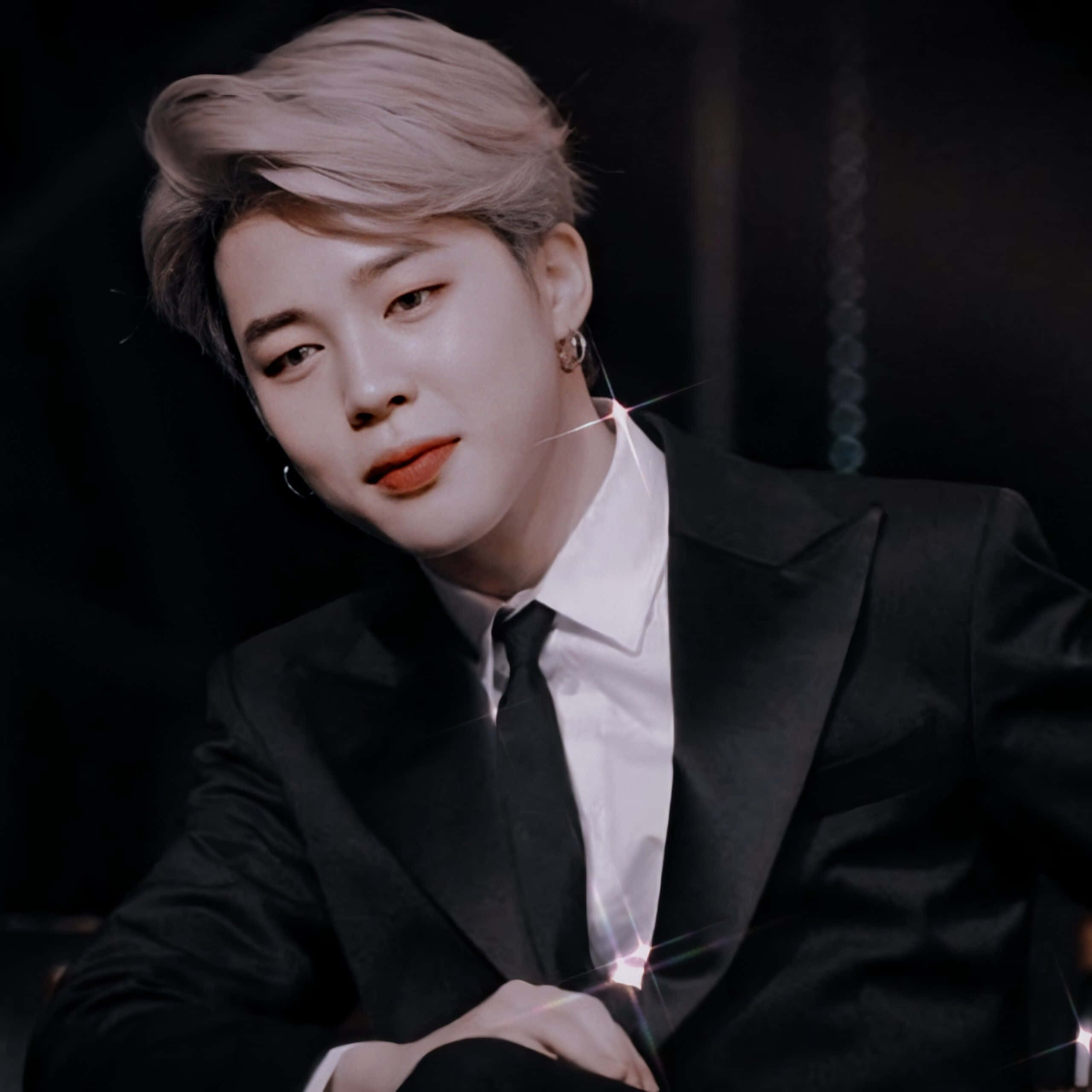 Jimin Aesthetic 桌布 壁紙 — 一位時尚男士，擁有鉑金色頭髮和紅唇，穿著黑色西裝，背景為深色並帶有鏡頭光暈。