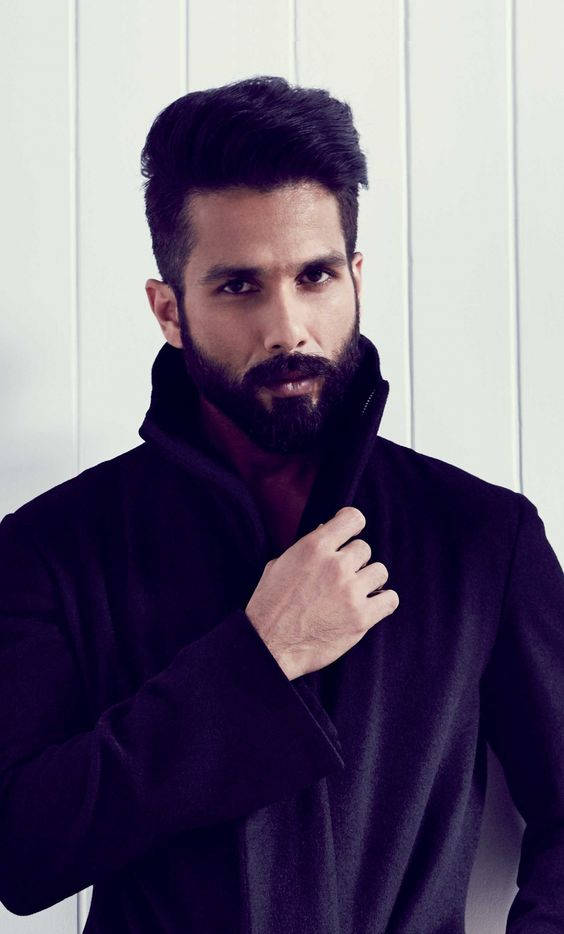 Shahid Kapoor 桌布 壁紙 — 一位時尚男士，留著濃密鬍鬚和深色頭髮，身穿黑色外套，靠著白色鑲板牆。