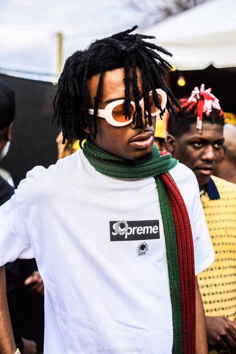Carti 桌布 壁紙 — 一位時尚男士，留著辮子頭，戴著白色太陽眼鏡，穿著Supreme T恤和綠色紅色圍巾，參加戶外活動。
