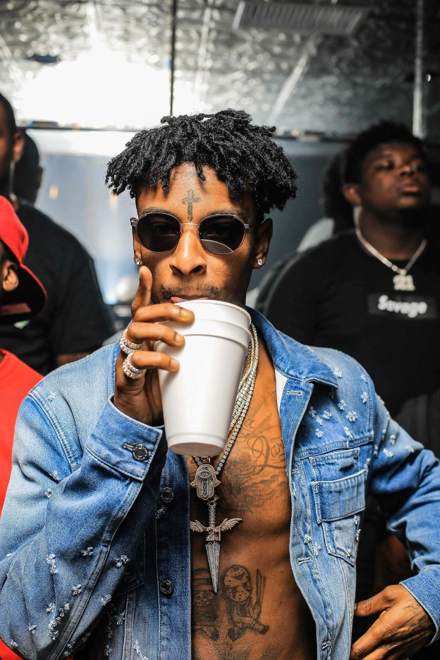 21 Savage 桌布 壁紙 — 一位時尚男士，留著辮子頭和刺青，戴著太陽眼鏡和牛仔外套，正從白色杯子中啜飲。