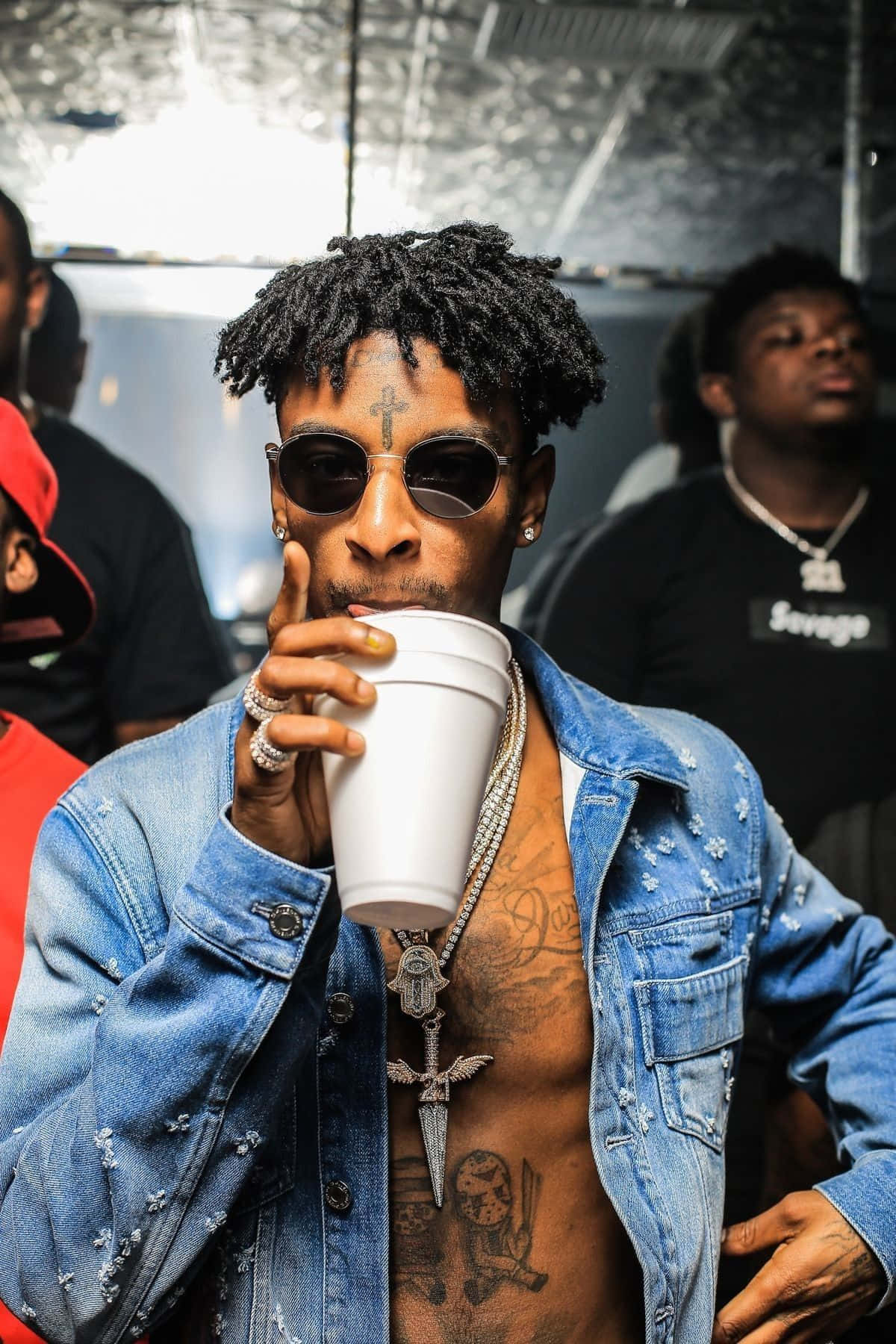 21 Savage 桌布 壁紙 — 一位時尚男士，留著辮子頭和刺青，戴著太陽眼鏡和牛仔外套，從白色杯子中啜飲。