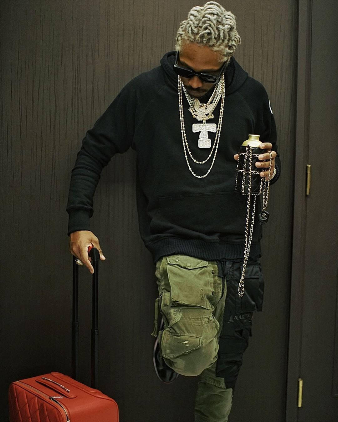 90s Rapper 桌布 壁紙 — 一位時尚男士，辮子頭髮和層疊鏈條，靠著深色牆壁，拿著瓶子，拉著紅色行李箱。