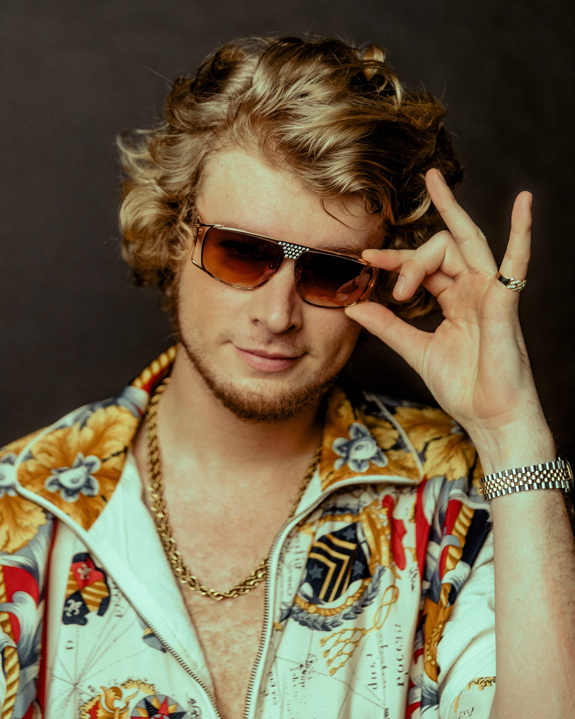 Sunglasses 桌布 壁紙 — 一位時尚男士，金色捲髮，戴著華麗太陽眼鏡和圖案外套，比出和平手勢。