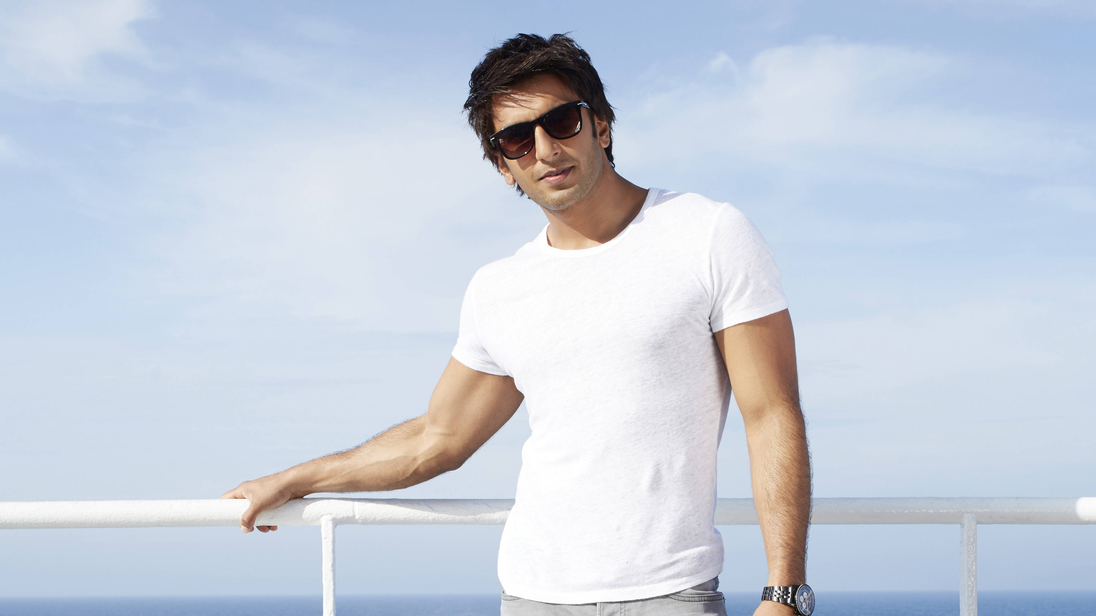 Ranveer Singh 桌布 壁紙 — 一位時尚男士戴著太陽眼鏡，隨意地靠在白色欄杆上，俯瞰著蔚藍天空下的海洋。