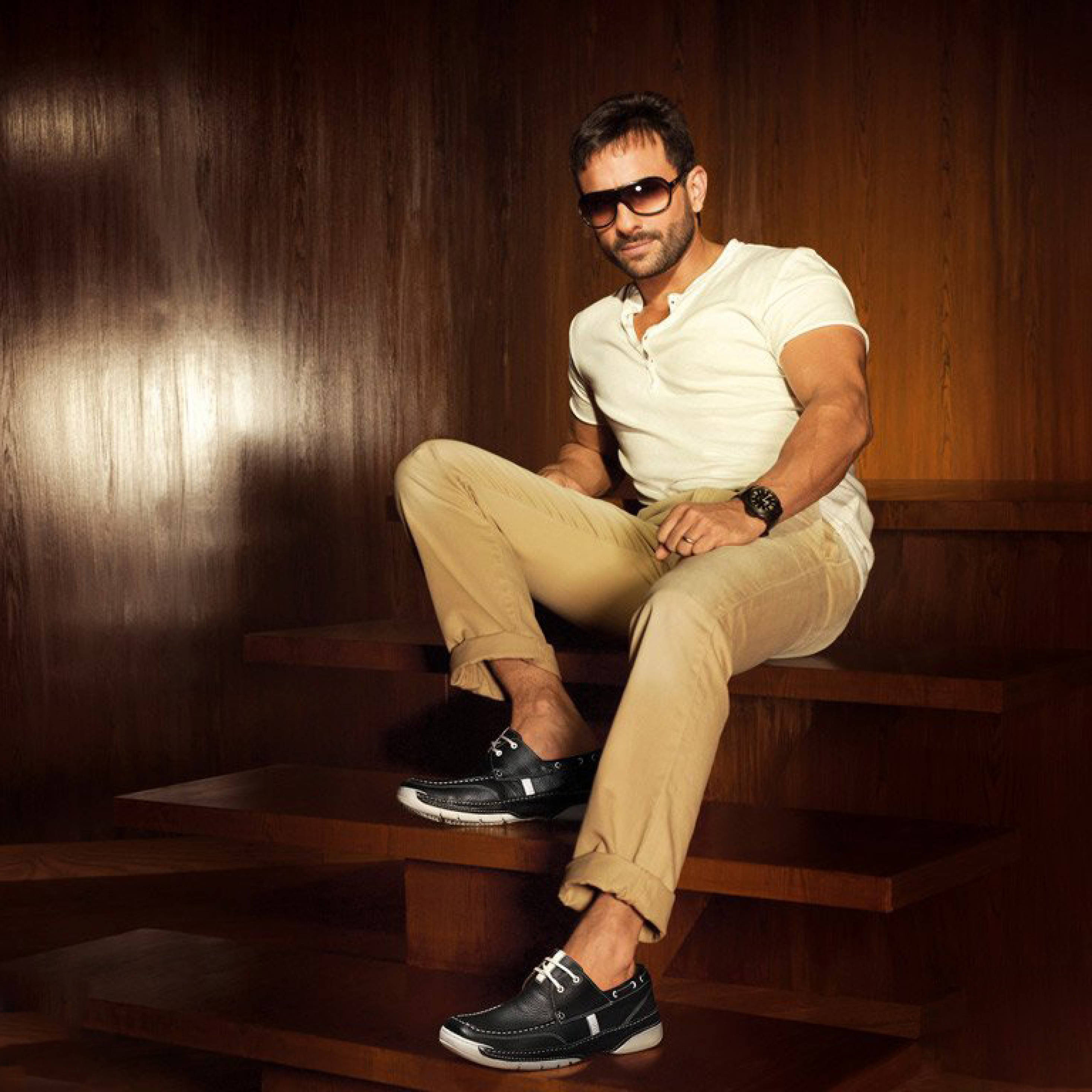 Saif Ali Khan 桌布 壁紙 — 一位時尚男士戴著太陽眼鏡和米色褲子，自信地坐在木階梯上，背景是溫暖的木鑲板牆。