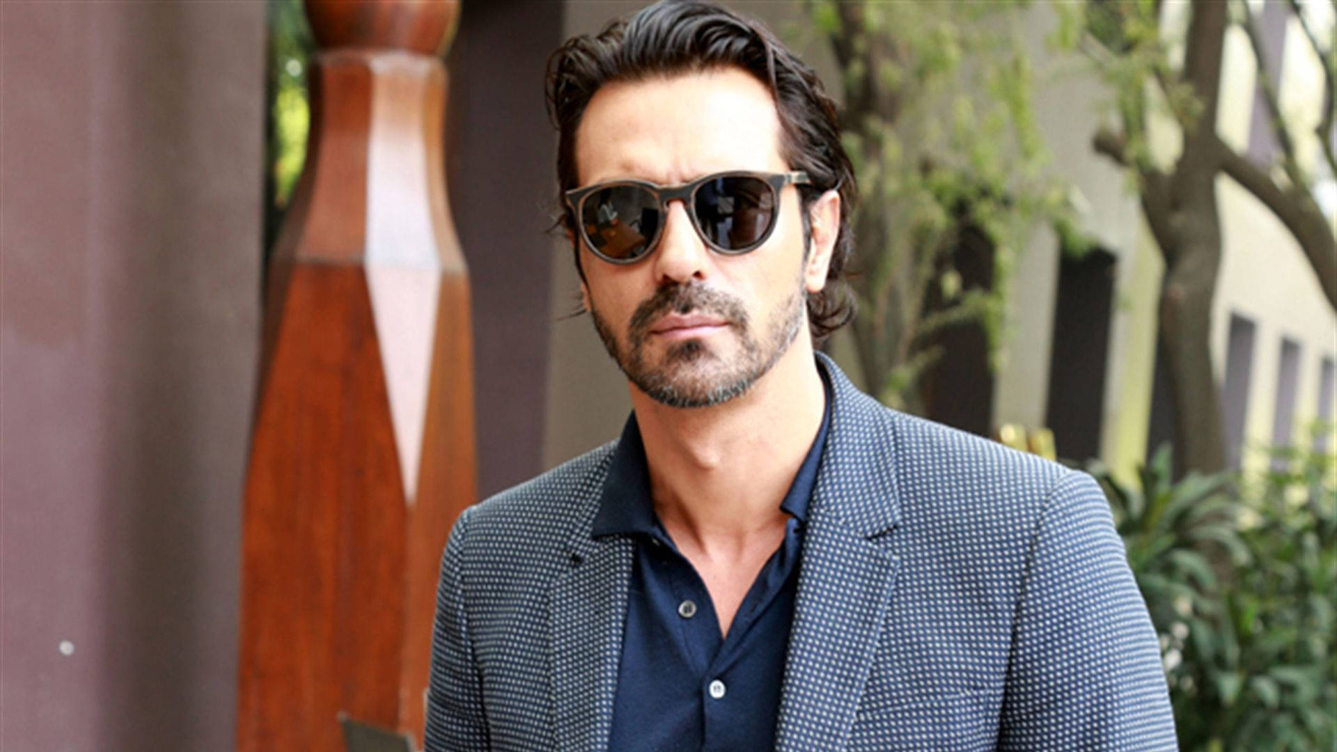 Arjun Rampal 桌布 壁紙 — 一位時尚男士戴著深色太陽眼鏡和圖案西裝外套，自信地站在戶外，靠近綠植和木雕。