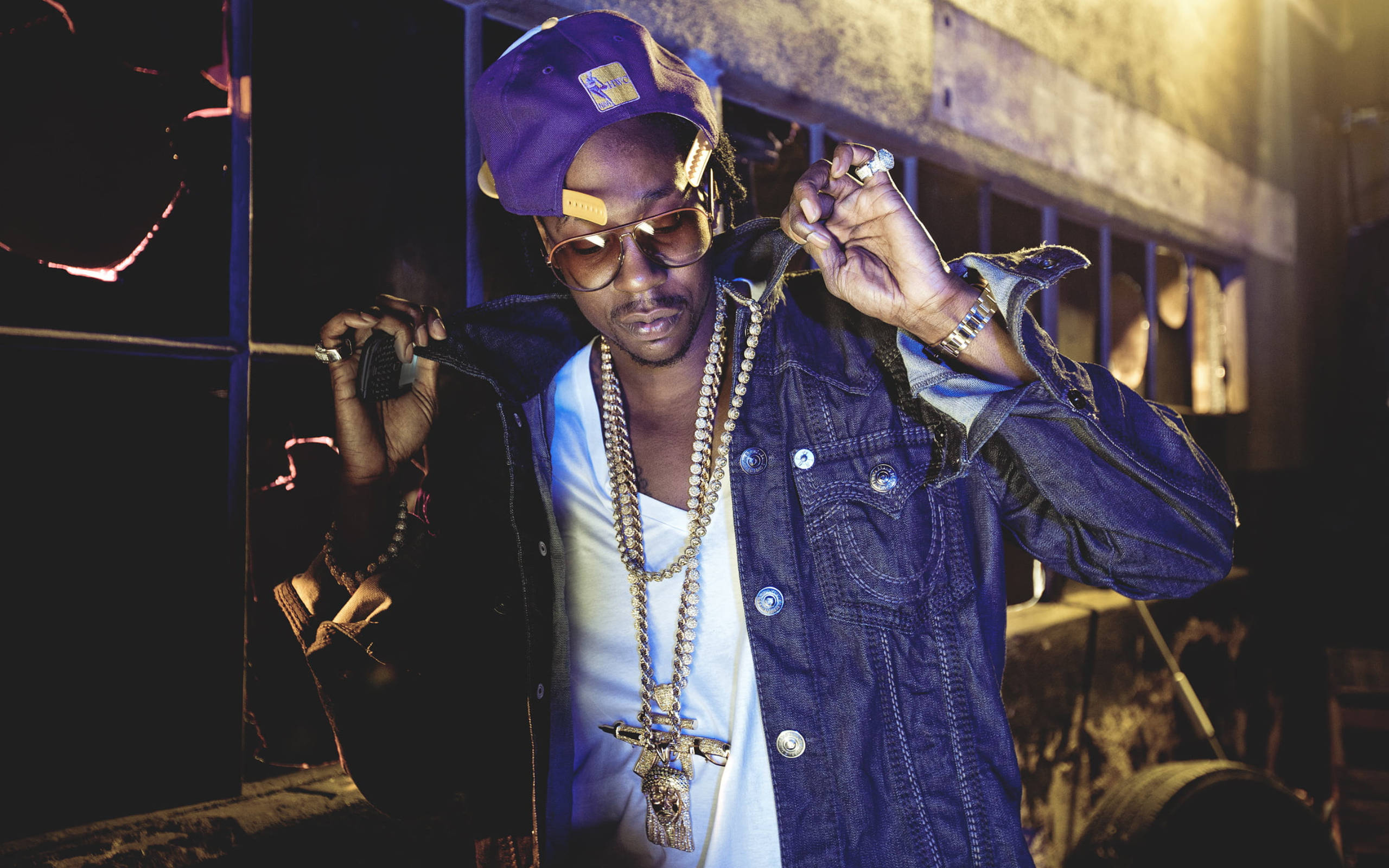 2 Chainz 桌布 壁紙 — 一位時尚男士戴著紫色帽子和牛仔外套，在溫暖的夜間燈光下自信地佩戴金鏈擺姿勢。