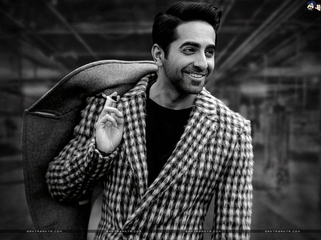 ayushmann khurrana 桌布 壁紙 — 一位時尚男士穿著千鳥格大衣，在模糊的城市背景下微笑著拿著香煙。