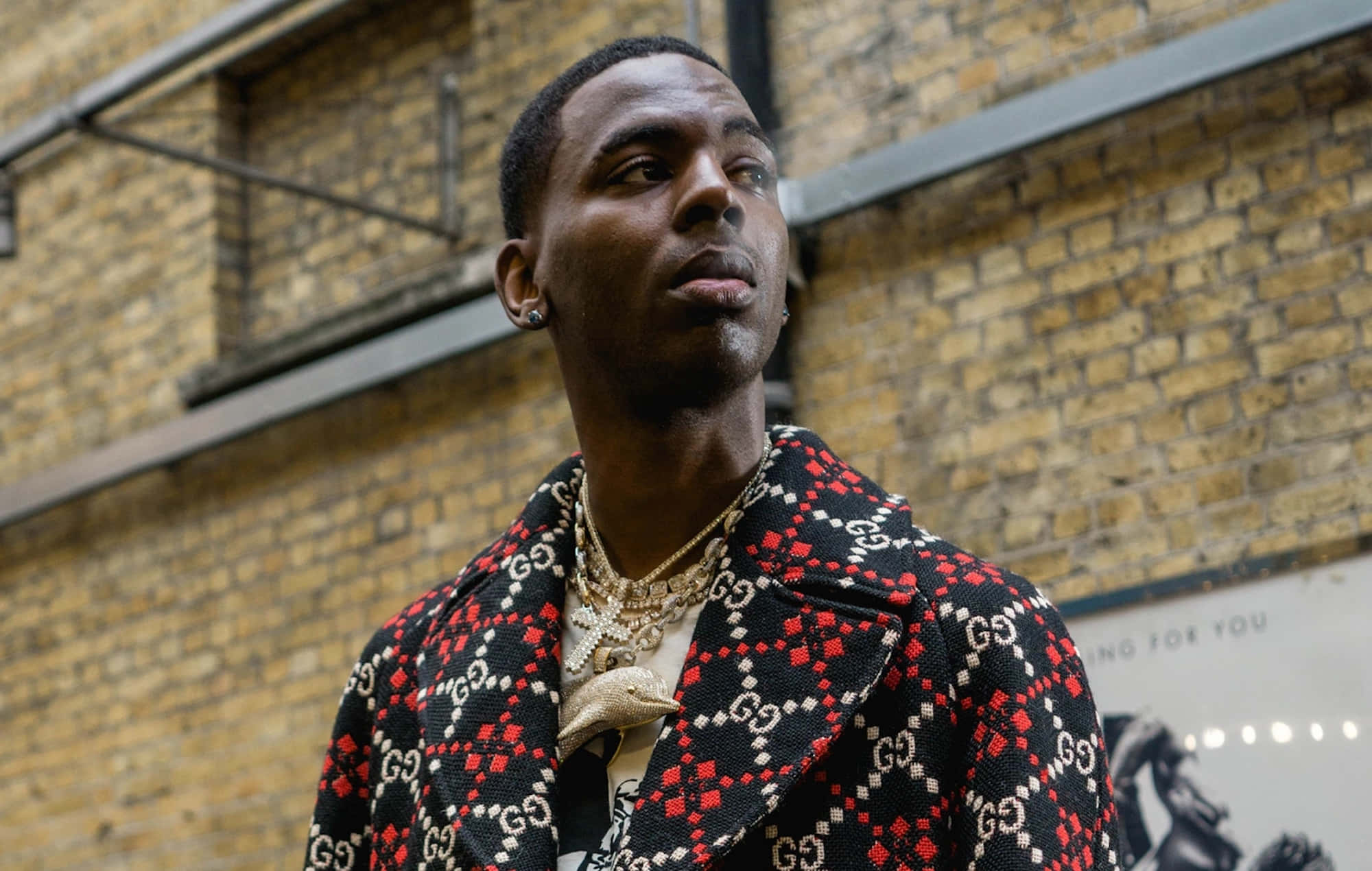 Young Dolph 桌布 壁紙 — 一位時尚男士穿著大膽的紅黑白三色Gucci圖案外套，站在風化的磚牆前。