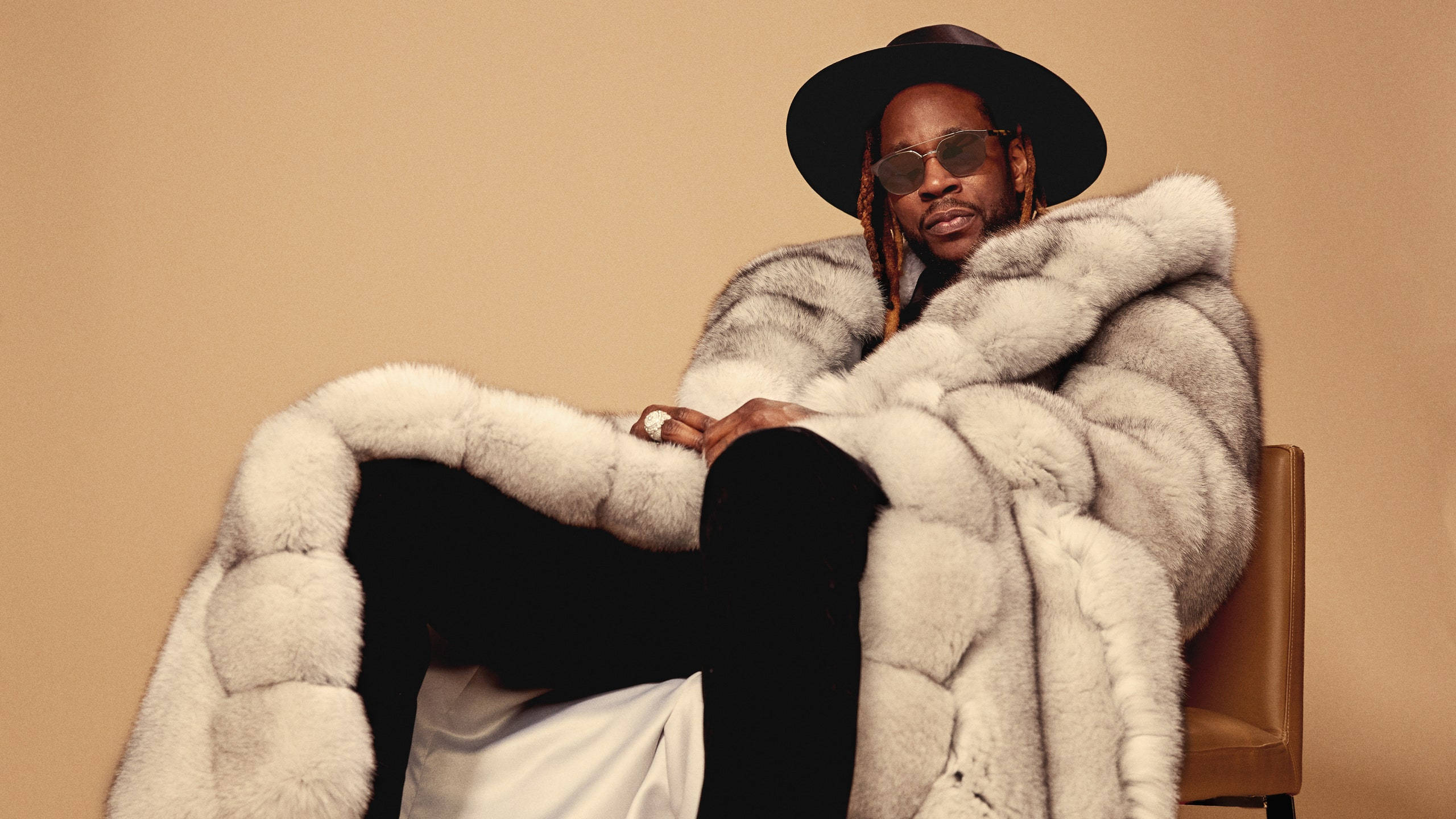 2 Chainz 桌布 壁紙 — 一位時尚男士穿著寬鬆的銀色毛皮大衣和黑色帽子，自信地靠在溫暖米色背景的椅子上。