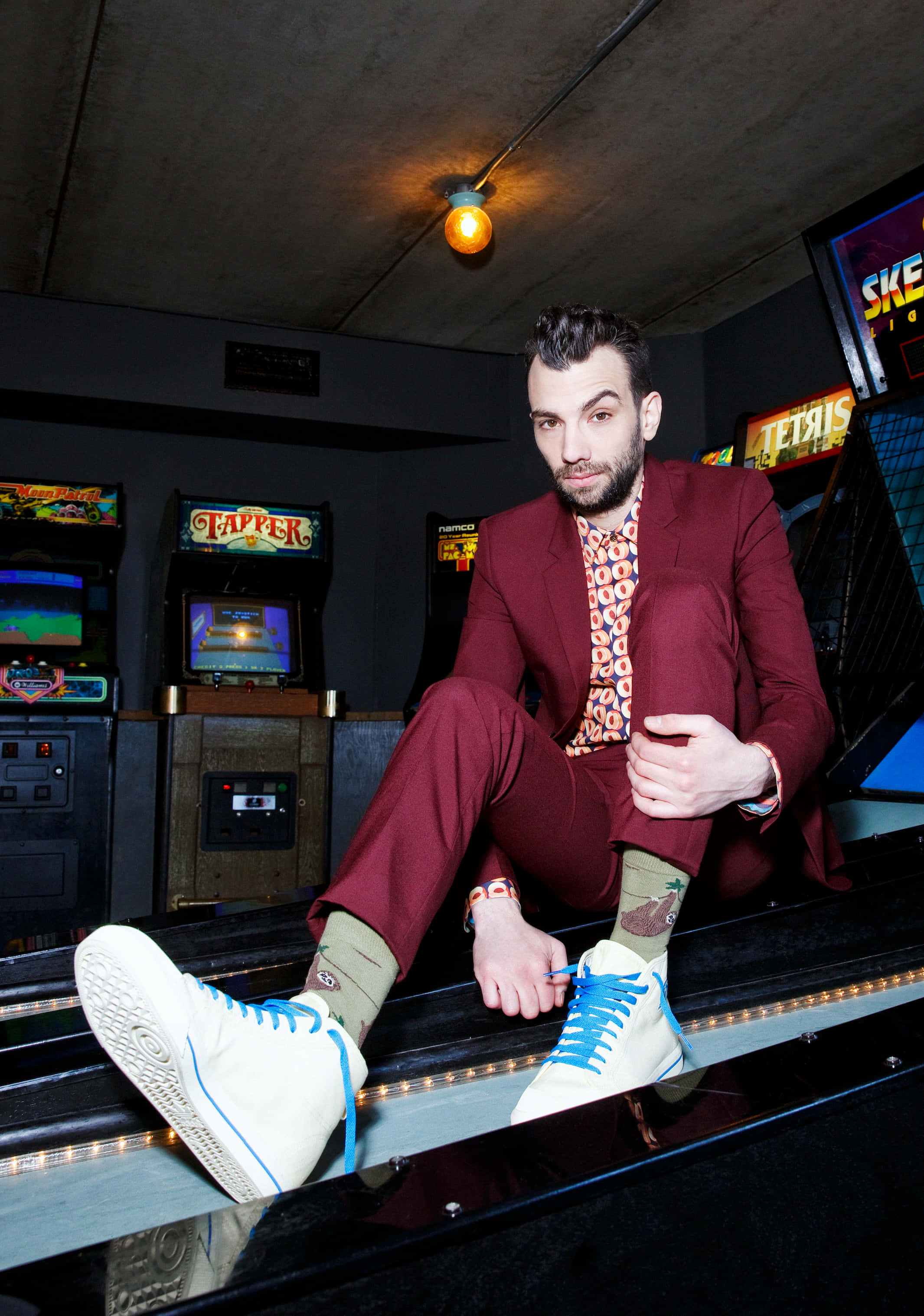 Jay Baruchel 桌布 壁紙 — 一位時尚男士穿著栗色西裝和白色運動鞋，自信地坐在昏暗復古遊戲室中的一台懷舊街機上。